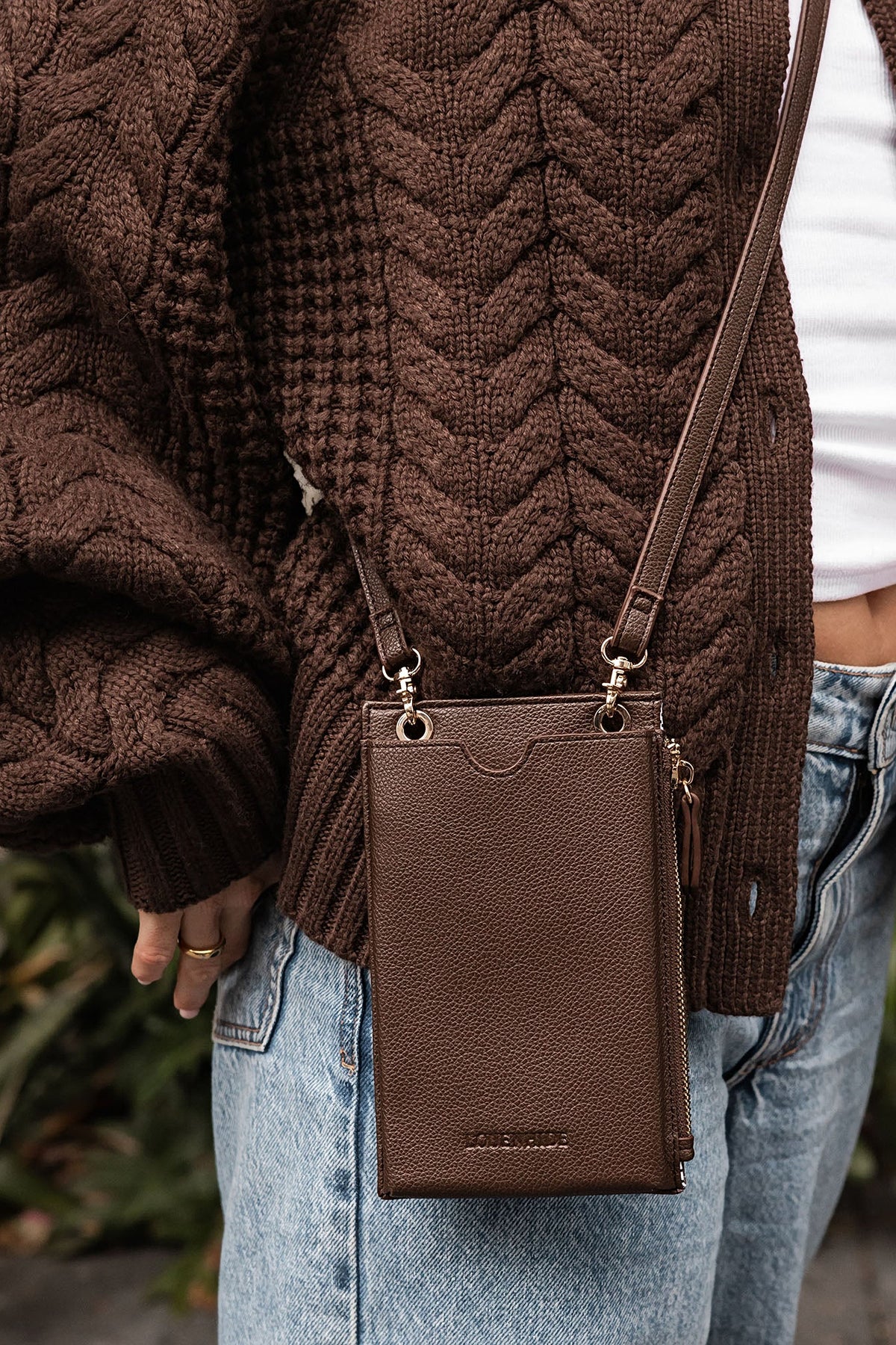 The Chocolate Suki Phone Crossbody Bag worn across the body at the hip with a brown cable knit cardigan and jeans.
