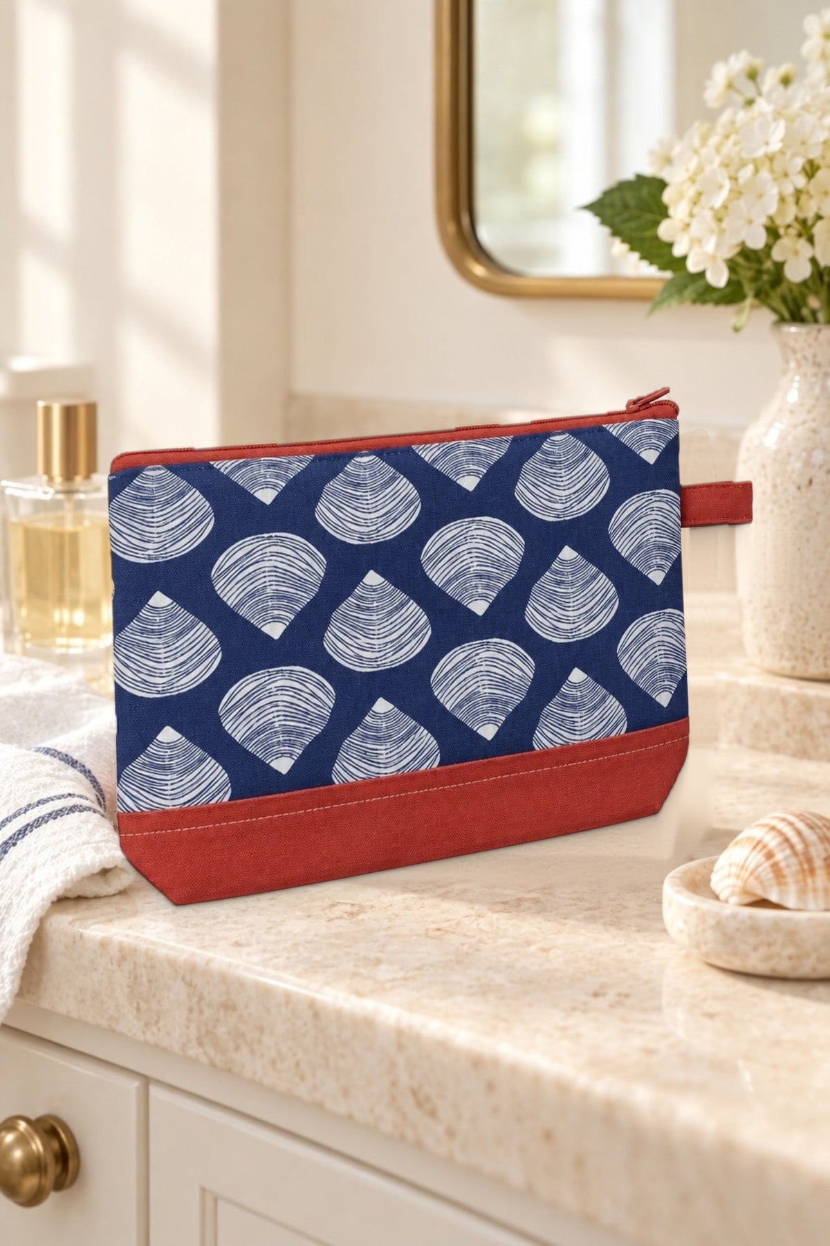 The Clamshell cosmetic bag on a bathroom vanity beside a mirror and white flowers.