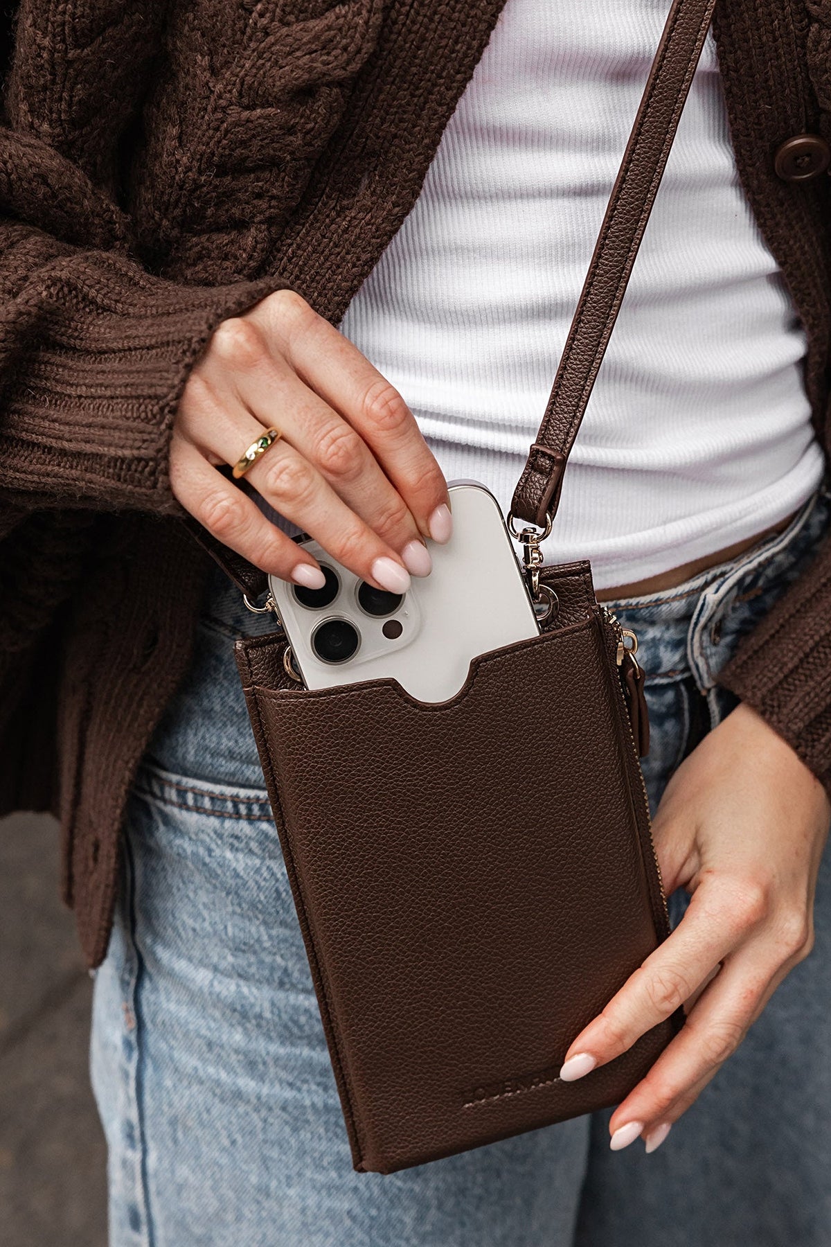 A hand sliding a phone into the Chocolate Suki Phone Crossbody Bag worn with a white tank, a brown cardigan and jeans.