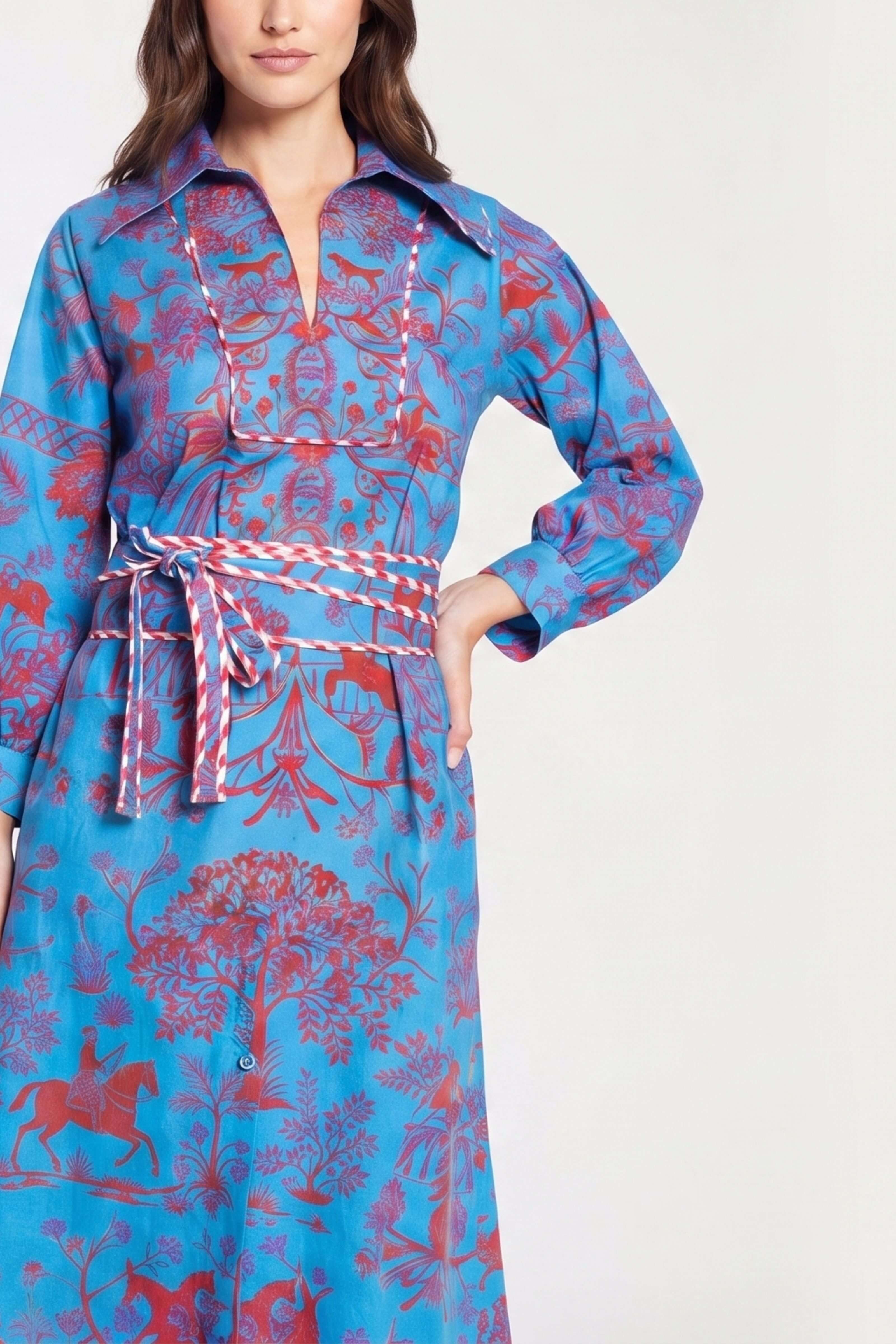 A woman wearing a long-sleeve collared dress with a blue base and red botanical and animal print, featuring a matching red-and-white patterned tie belt at the waist.