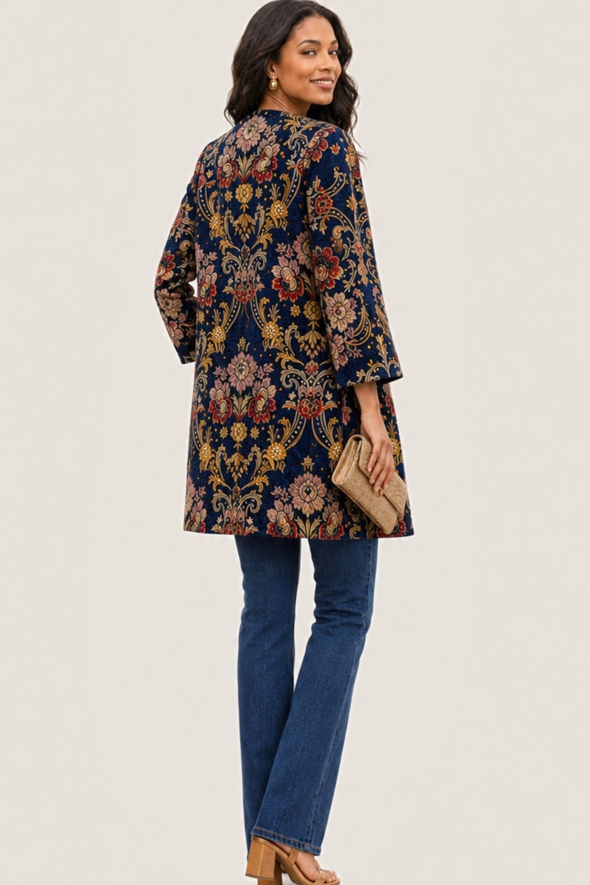 A woman modeling a navy blue tunic top with an intricate multicolor floral and paisley pattern, paired with blue jeans and holding a beige clutch bag against a plain off-white background.