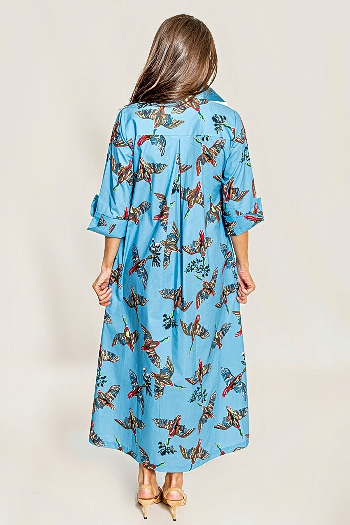 A woman shown from the back blue maxi dress with birds print wstanding against a plain beige studio background.