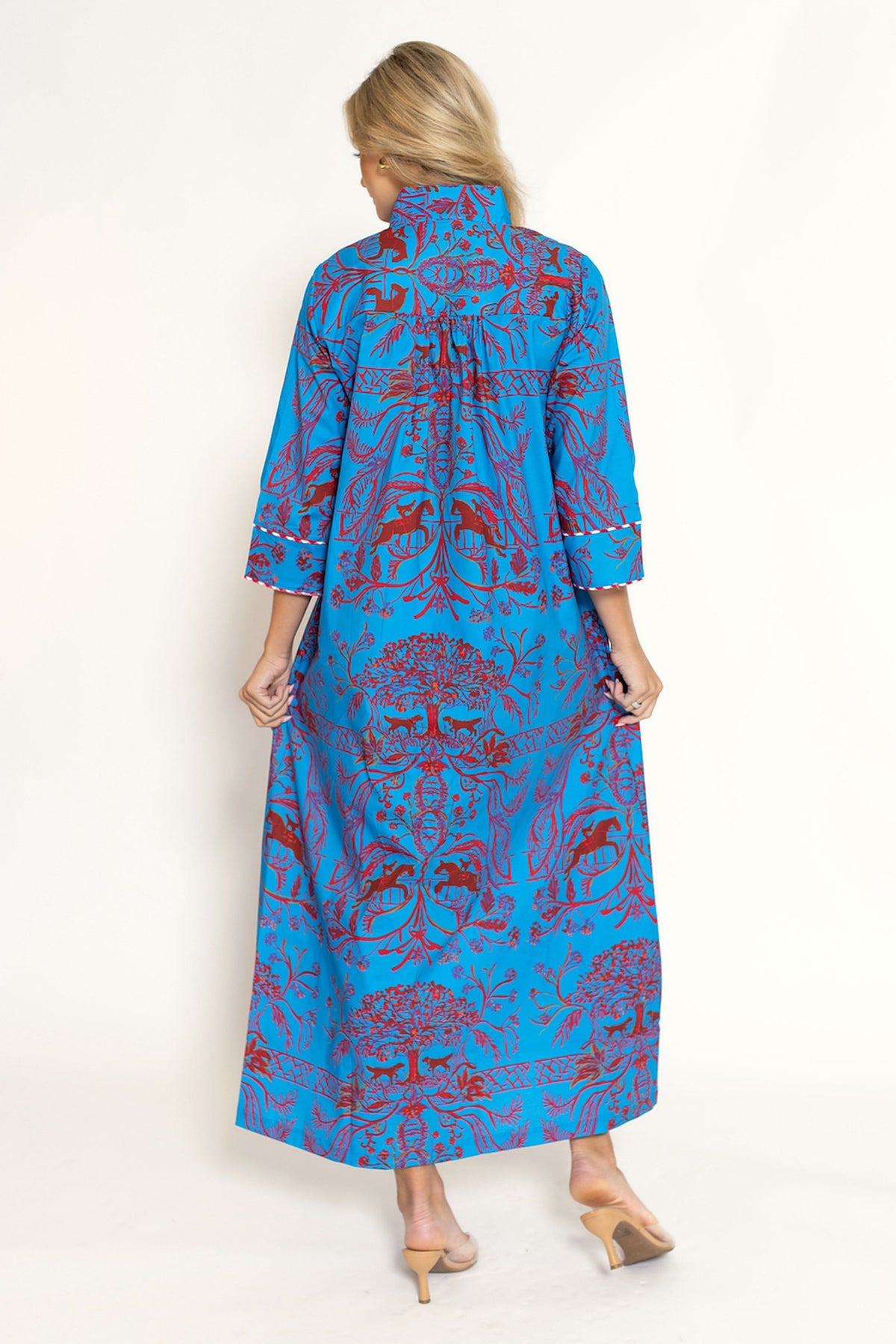 A woman viewed from the back wearing a long blue dress with a red and purple botanical pattern, standing against a plain off-white studio background.