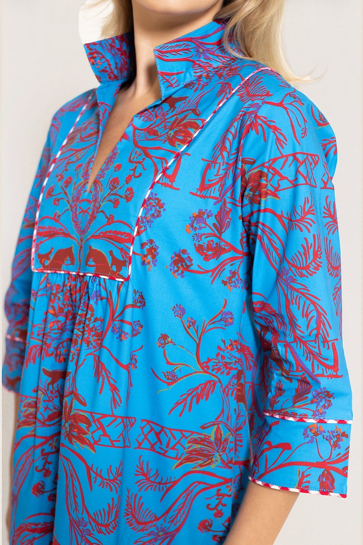 A close-up of a woman wearing a bright blue shirt dress with a red botanical and floral print, featuring white contrast stitching along the collar and cuffs.