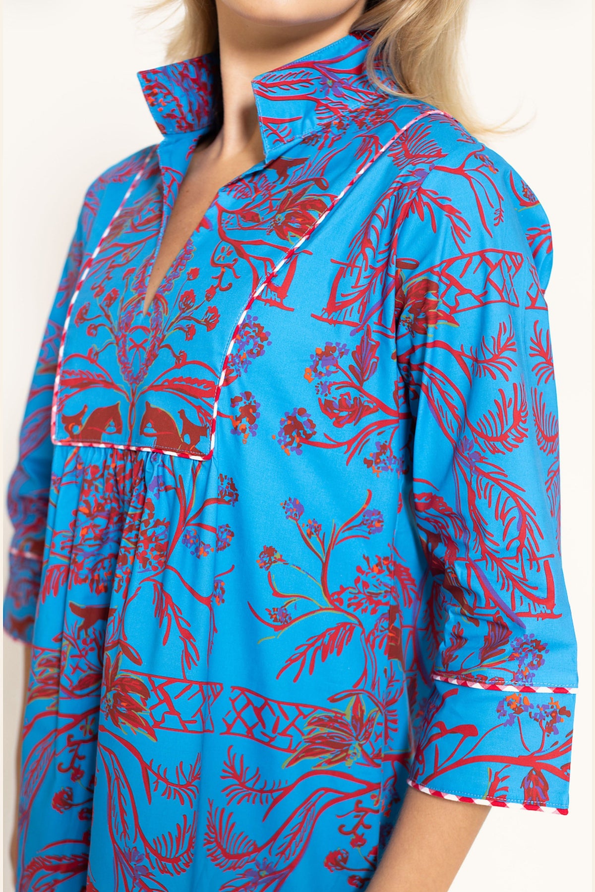 A close-up of a woman wearing a bright blue shirt dress with a red botanical and floral print, featuring white contrast stitching along the collar and cuffs.