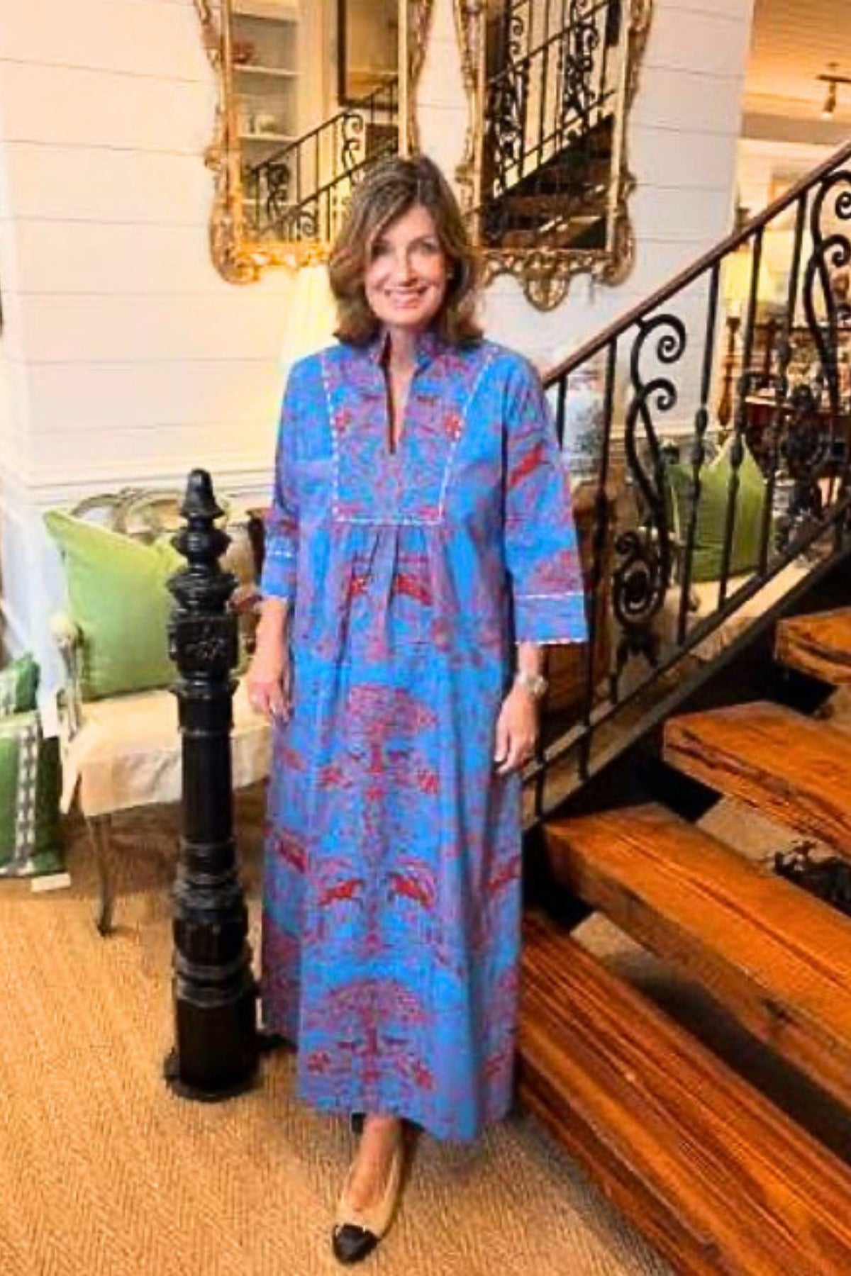 A woman wearing a blue and red patterned maxi dress stands in a home interior next to a wooden staircase.