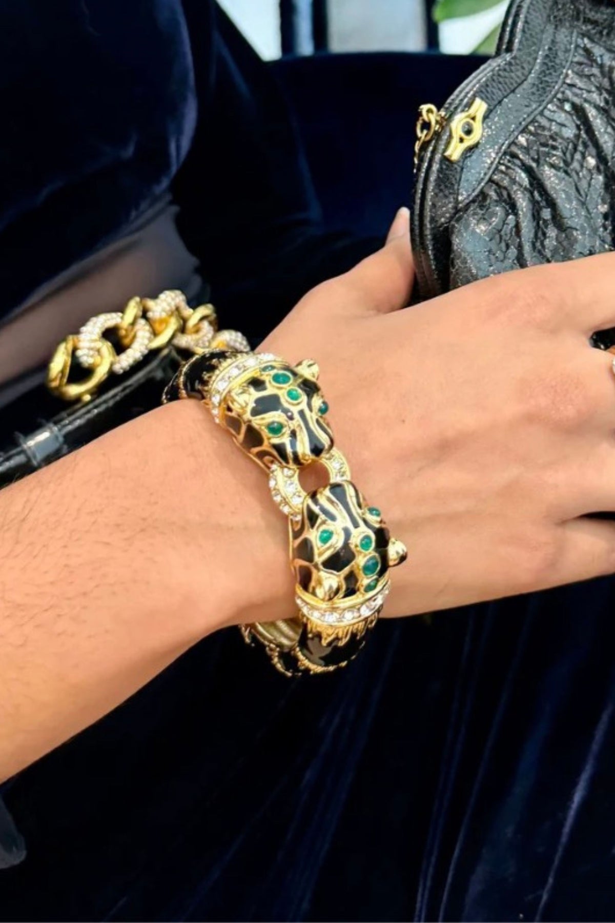 A close-up of a person's wrist wearing a chunky gold-toned bracelet with black enamel, green stone accents, and rhinestone details, alongside a black textured handbag.