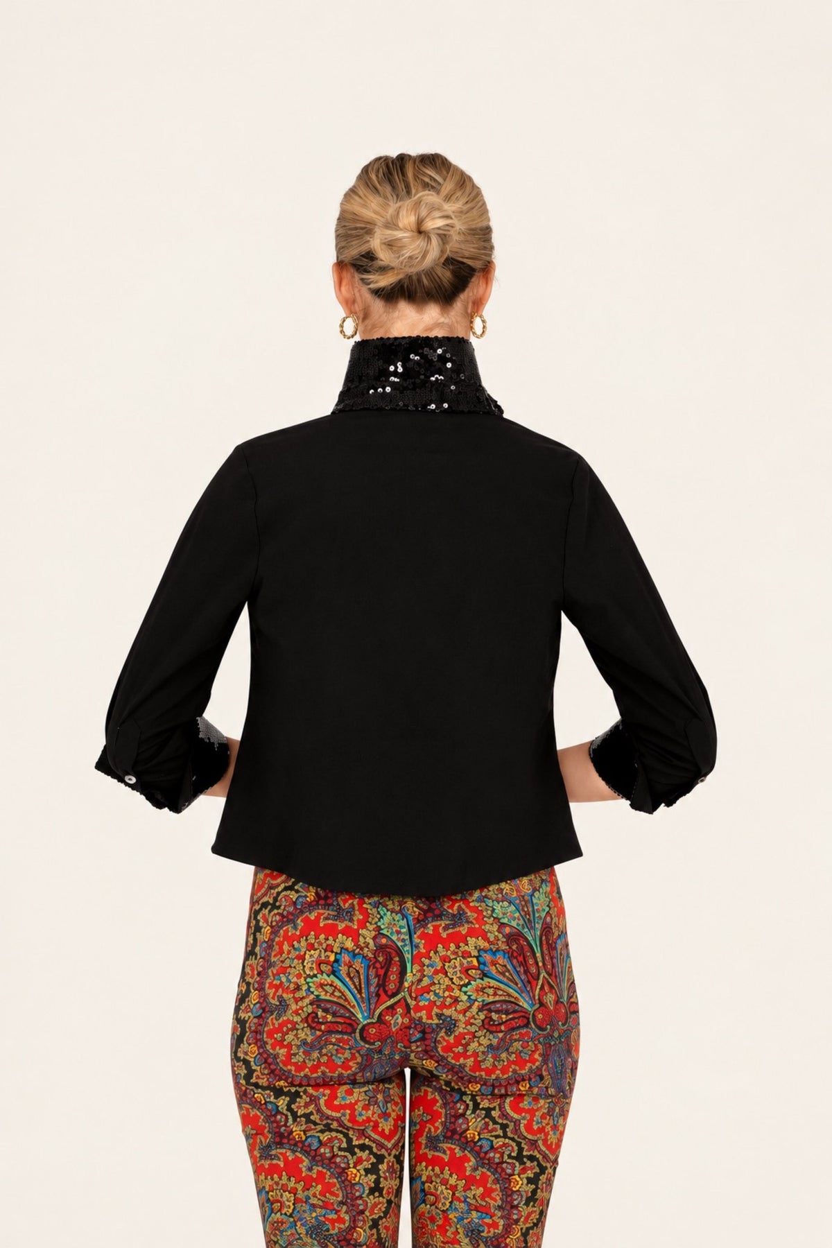 A woman shown from the back wearing a black cropped long-sleeve top with a high embellished collar and colorful paisley-print pants, against a plain light background.
