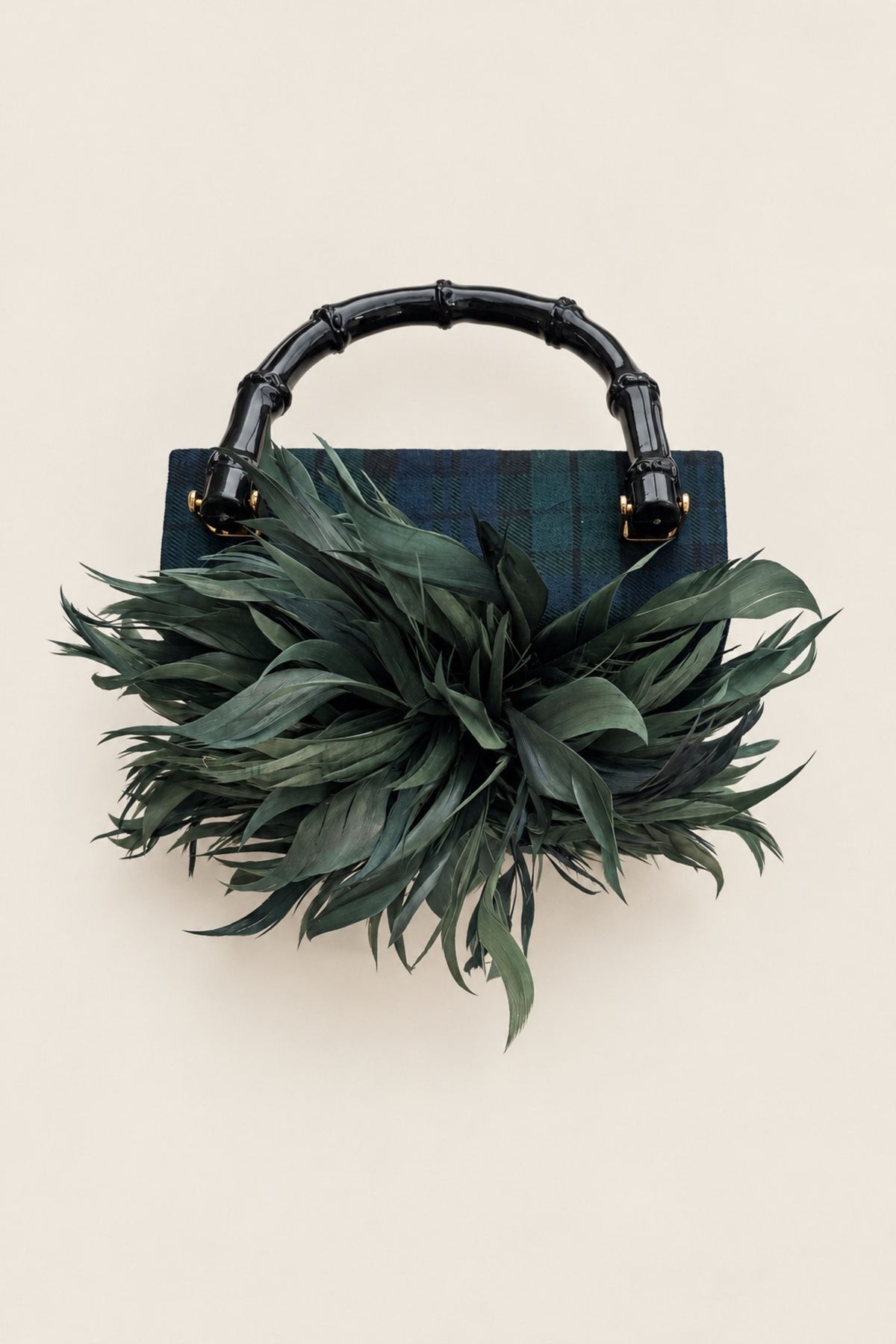 A structured handbag with a dark green plaid body, a glossy black bamboo-style top handle, and a large, voluminous dark green feather or leafy embellishment in front, photographed against a plain warm beige background.
