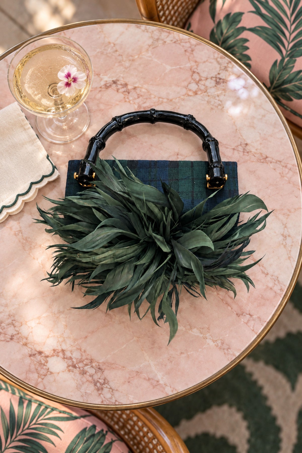 A green handbag with a bamboo-style top handle and a large green feather or leafy embellishment in front, displayed on a round pink marble table with a glass and tropical-patterned chair visible.