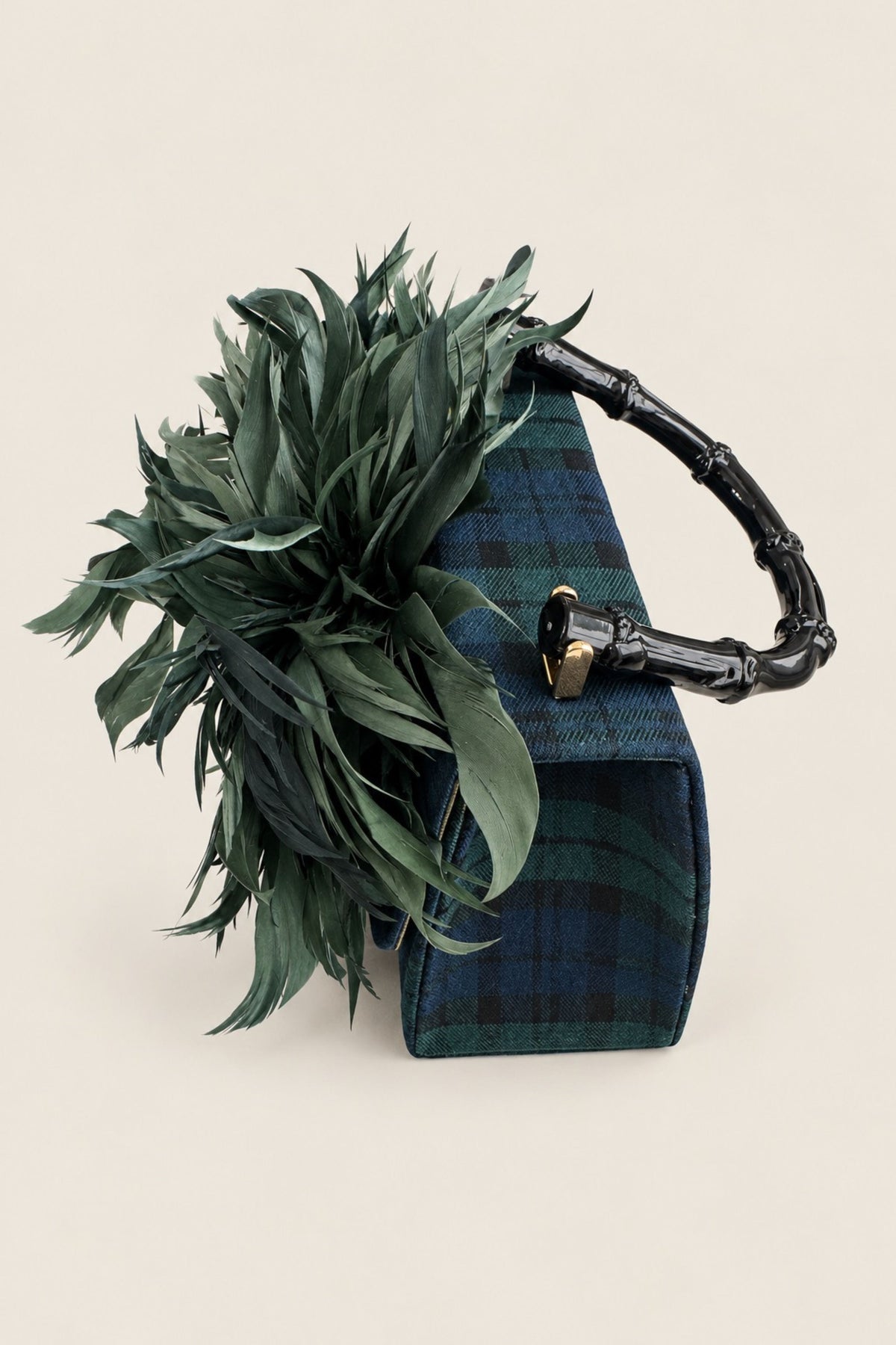 A structured handbag with a dark green and blue plaid pattern, a black bamboo-style handle, and a large green feather embellishment on the front, set against a solid light beige background.