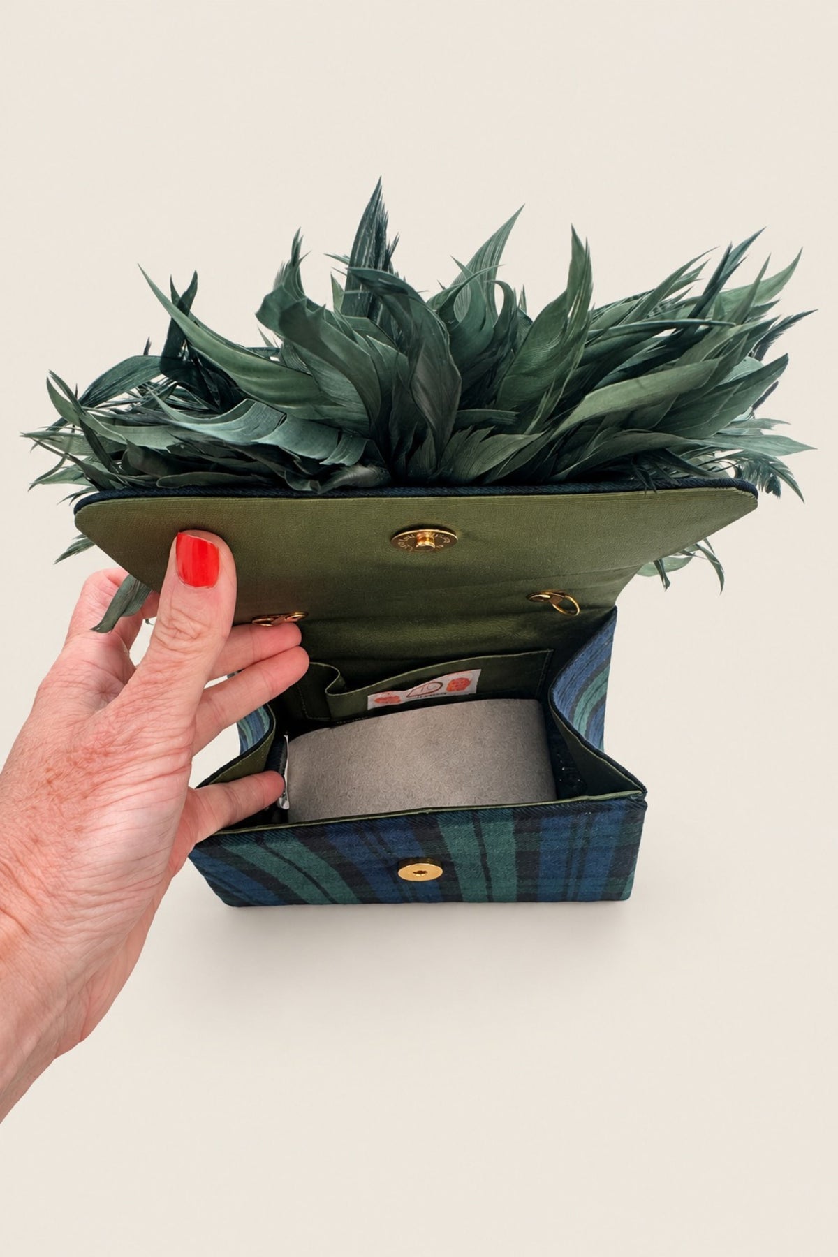 A hand with red nail polish holds open a small green and blue plaid clutch bag with gold hardware, revealing a gray interior and a green leafy arrangement on top, against a plain beige background.