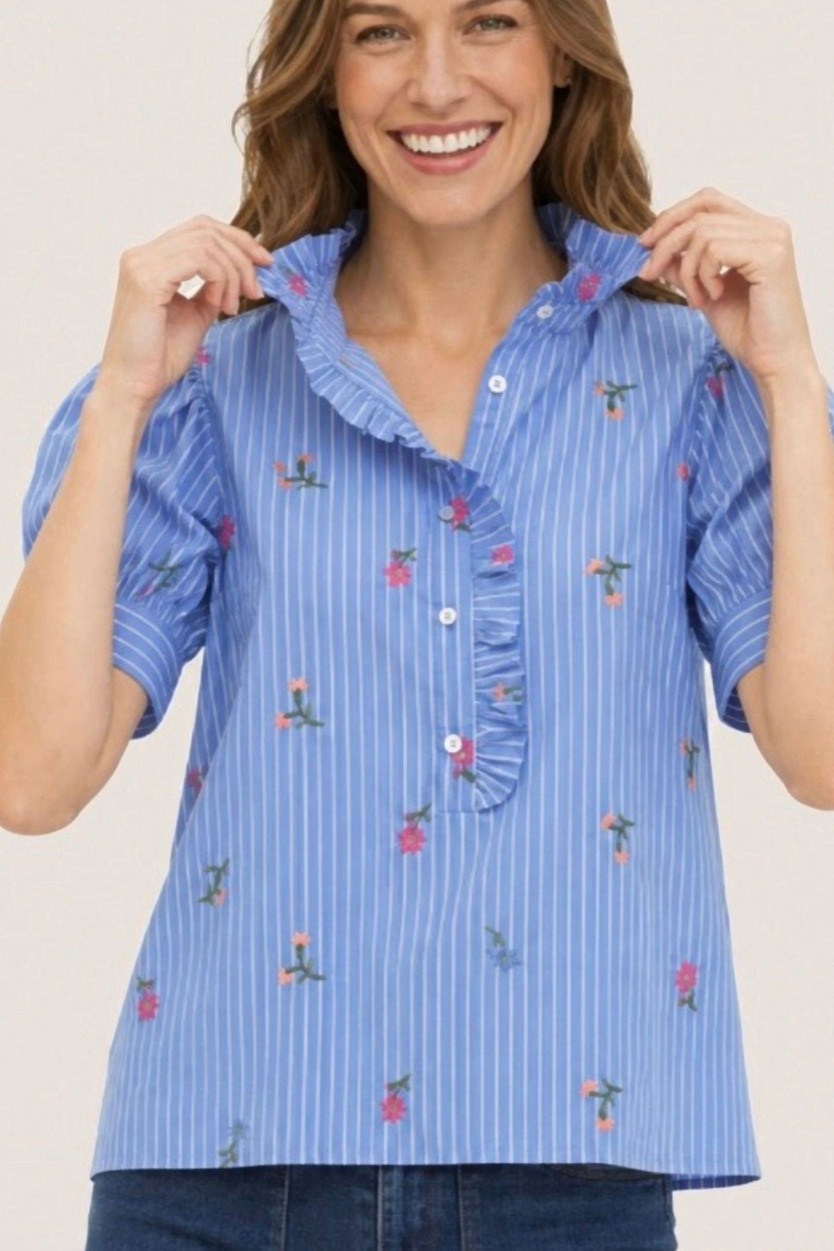 A woman wearing a light blue and white vertically striped short-sleeve blouse with ruffled trim and small pink floral embroidery, paired with dark blue jeans against a plain white background.