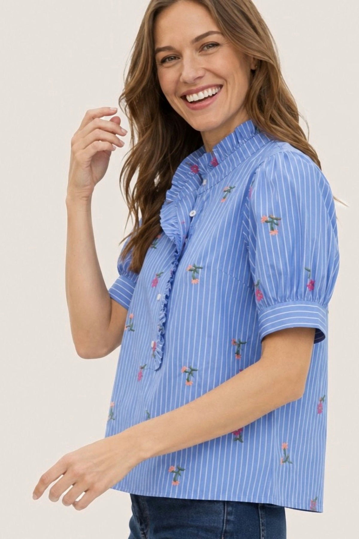 A woman wearing a light blue and white vertically striped short-sleeve blouse with small floral embroidery, paired with dark blue jeans, against a plain white background.