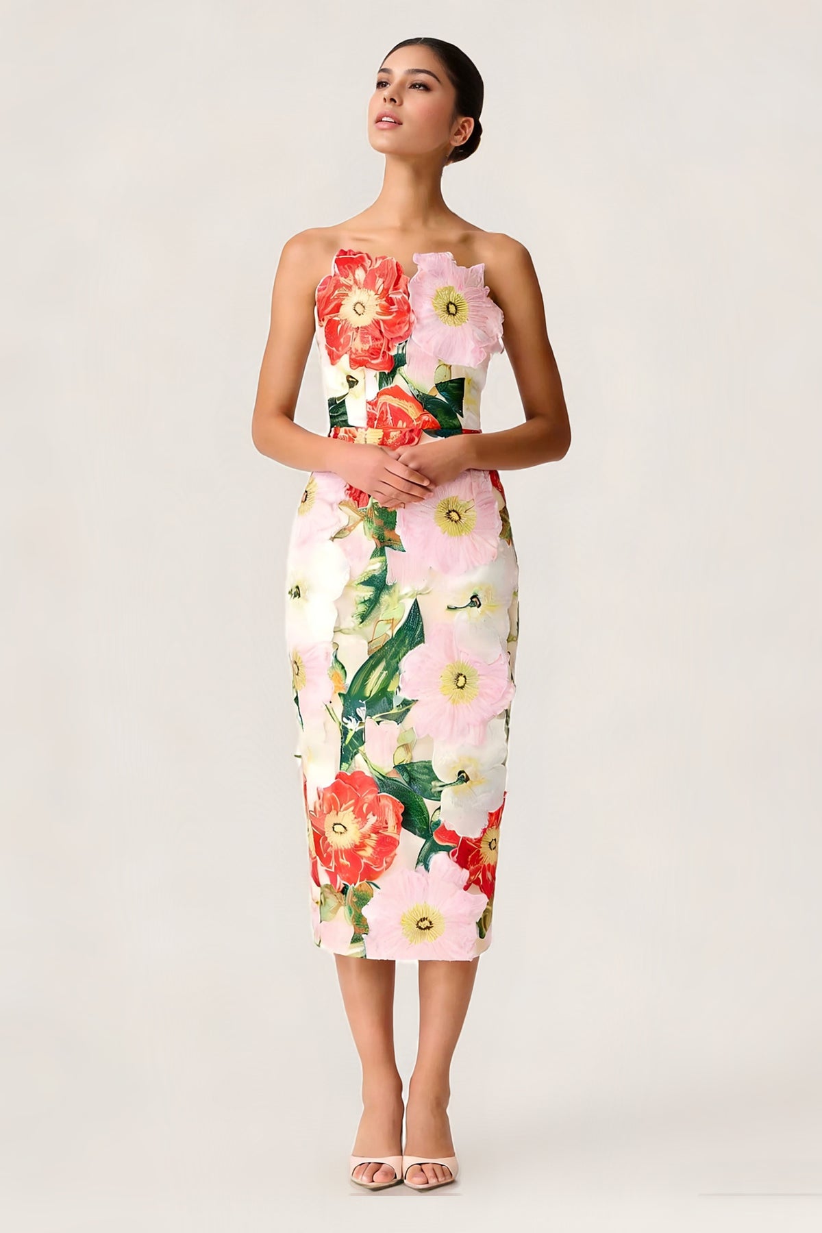 A woman modeling a strapless mini dress with a large 3D floral applique print in red, pink, white, and green on a cream background, paired with clear high-heeled sandals against a white studio backdrop.