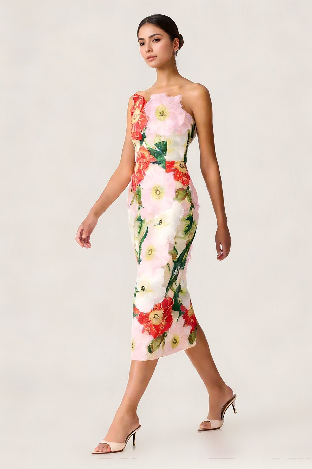 A woman models a one-shoulder midi dress featuring a vibrant 3D floral applique print on a light pink mesh base. The dress has large pink, red, and white flowers with green leaves, and she is wearing beige heeled sandals against a clean white studio background.