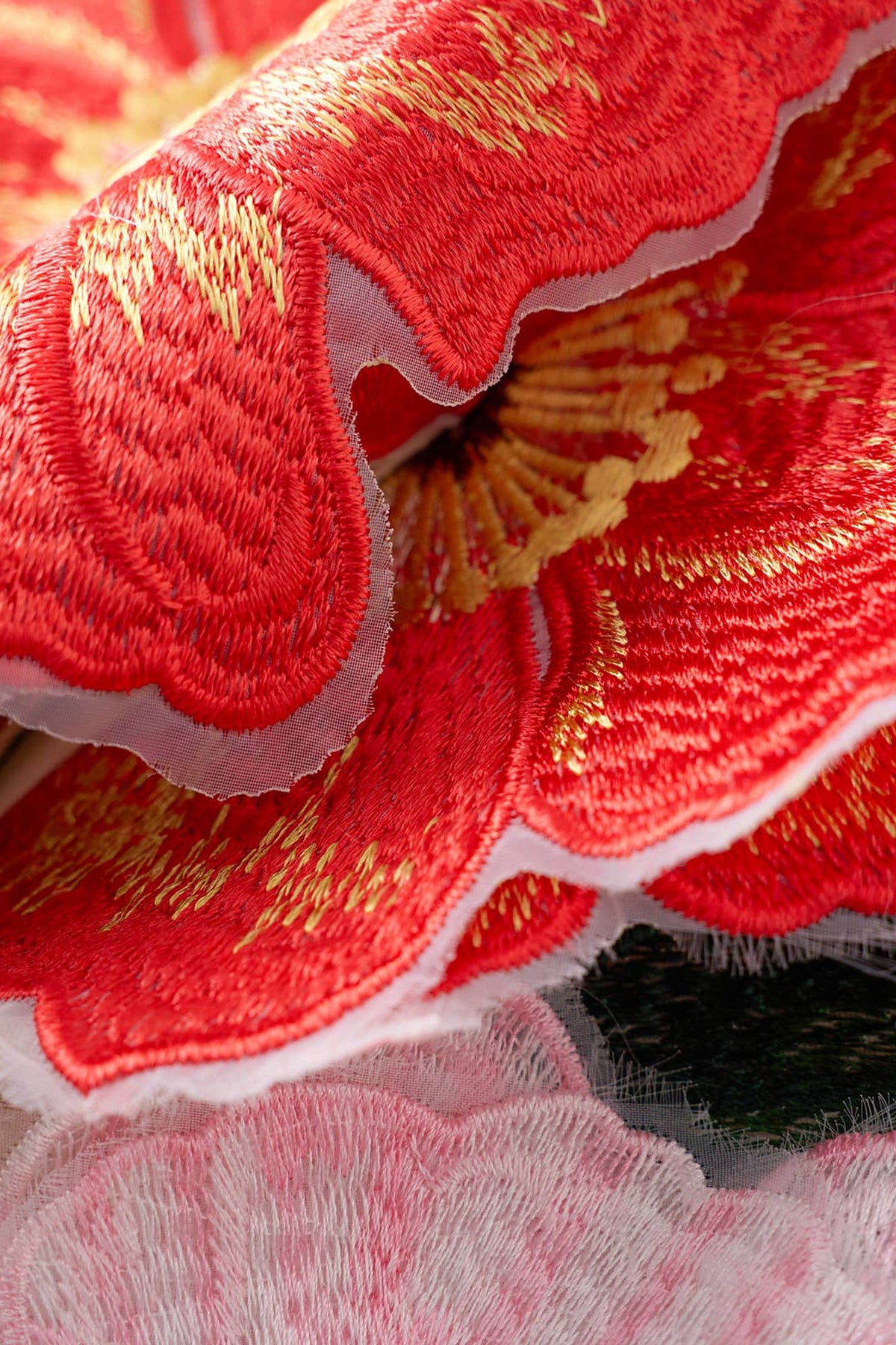A close-up of a red embroidered fabric flower with gold thread details and a sheer pink overlay.