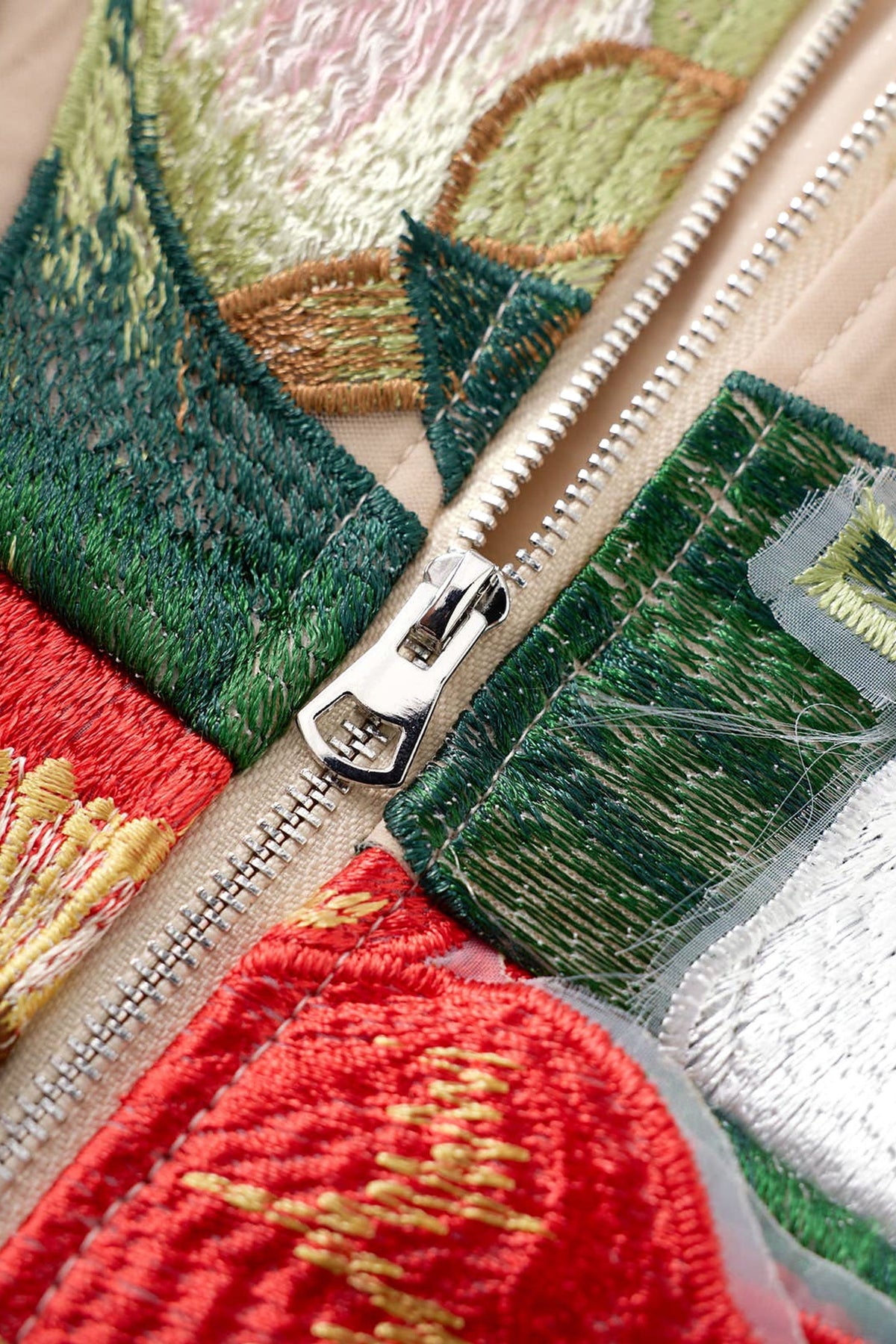 Close-up of a colorful embroidered zippered garment, showing green, red, beige, and gold embroidery with a silver metal zipper pull.