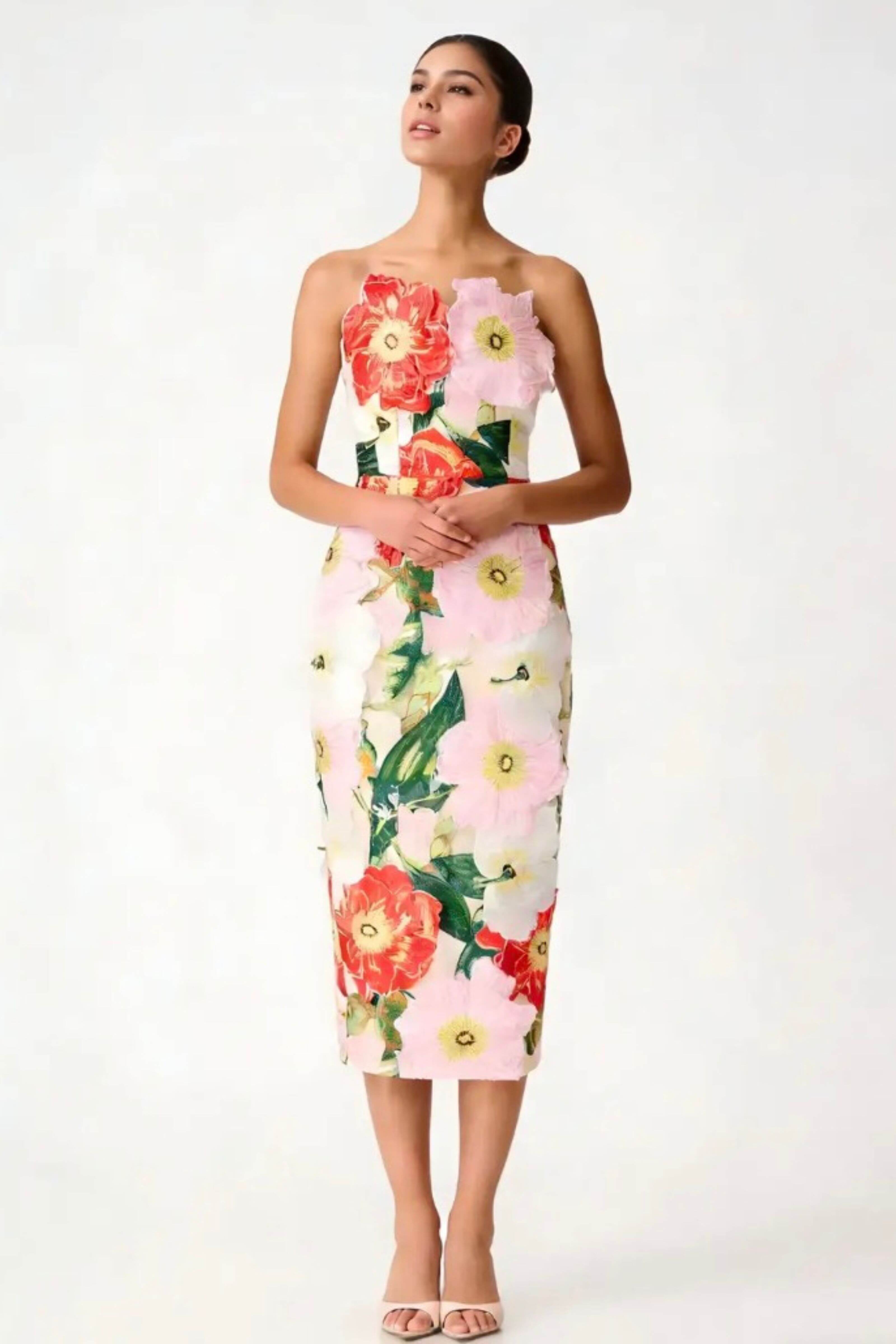 A woman modeling a strapless knee-length dress with a vibrant floral print featuring red, pink, and white flowers with green leaves on a light background, set against a plain white studio backdrop.