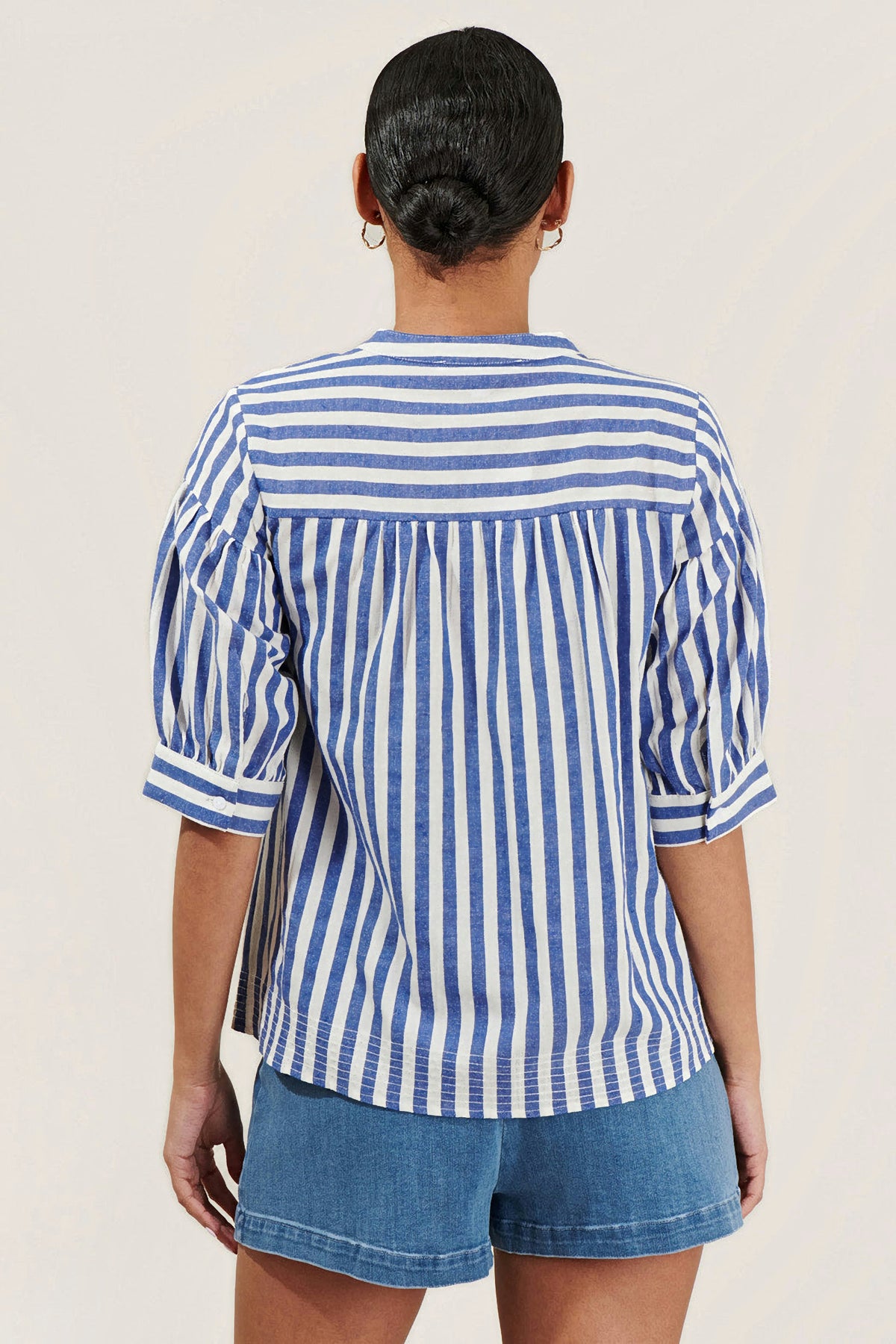 A woman viewed from the back wearing a blue and white vertically striped blouse with puff sleeves and blue jeans, set against a plain light beige background.