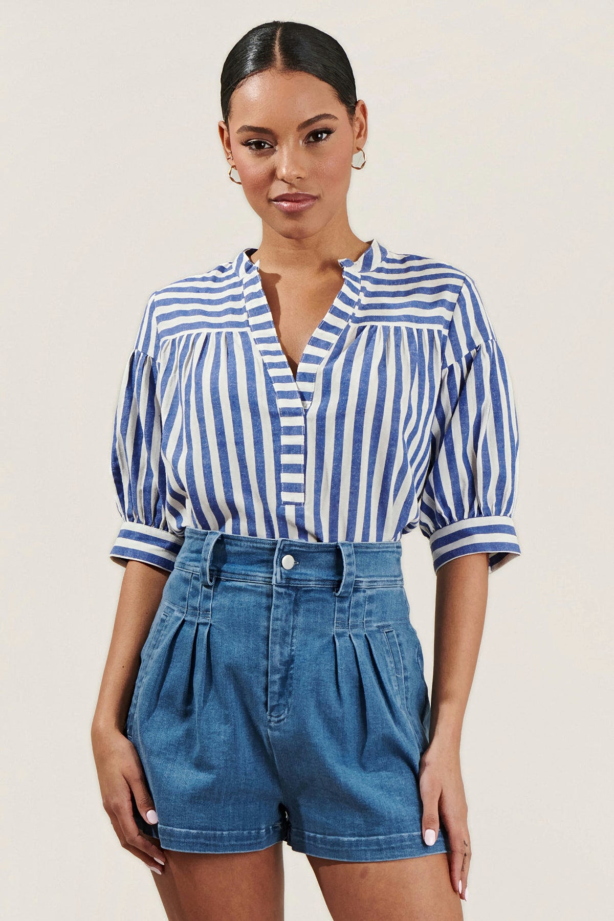 A woman wearing a blue and white vertically striped short-sleeve blouse with puff sleeves and a split neckline, paired with blue jeans, against a solid light beige background.