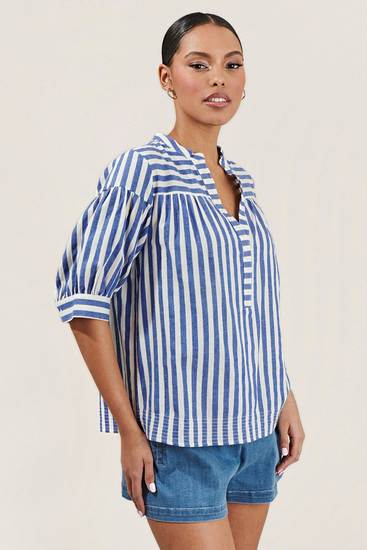 A woman wearing a blue and white vertically striped blouse with puff sleeves and a split neckline, paired with blue jeans, against a solid light beige background.