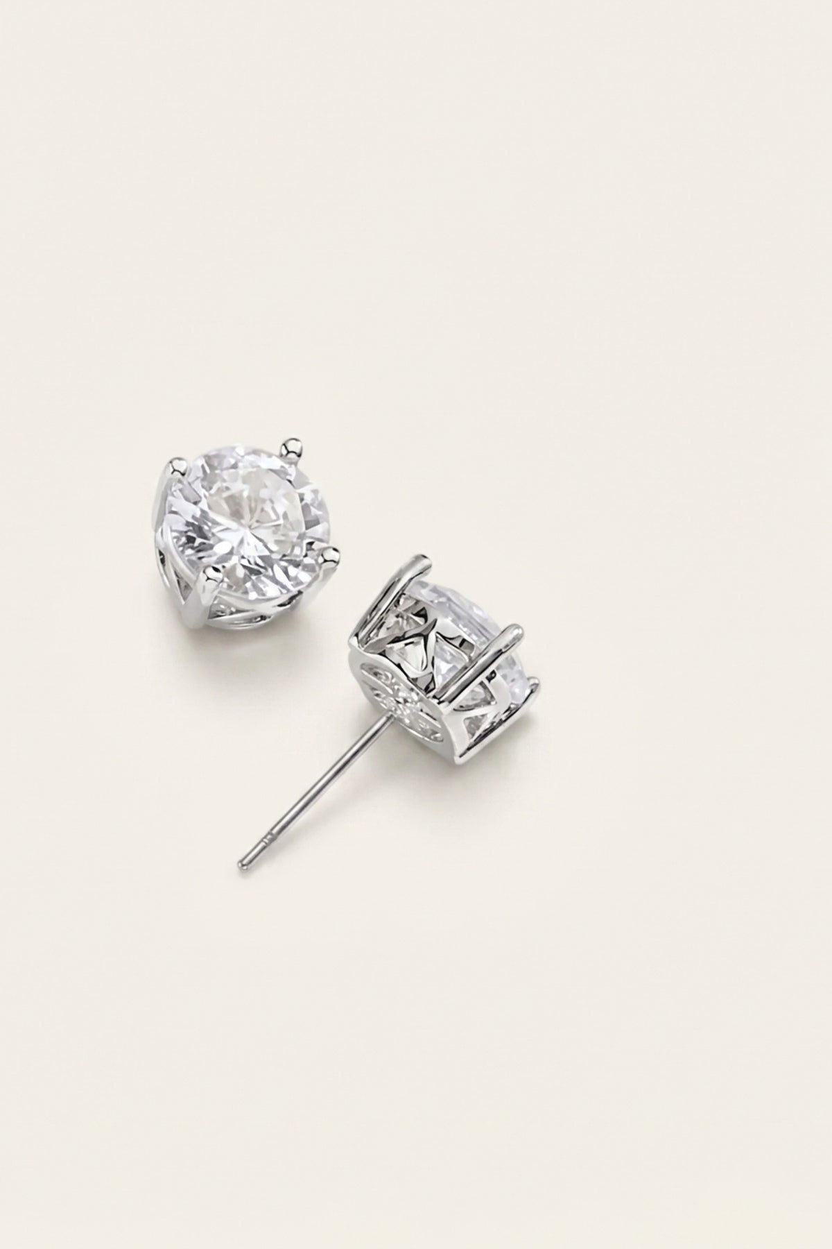 A pair of silver stud earrings with clear gemstones, one shown from the front and the other from the side, on a light beige background.