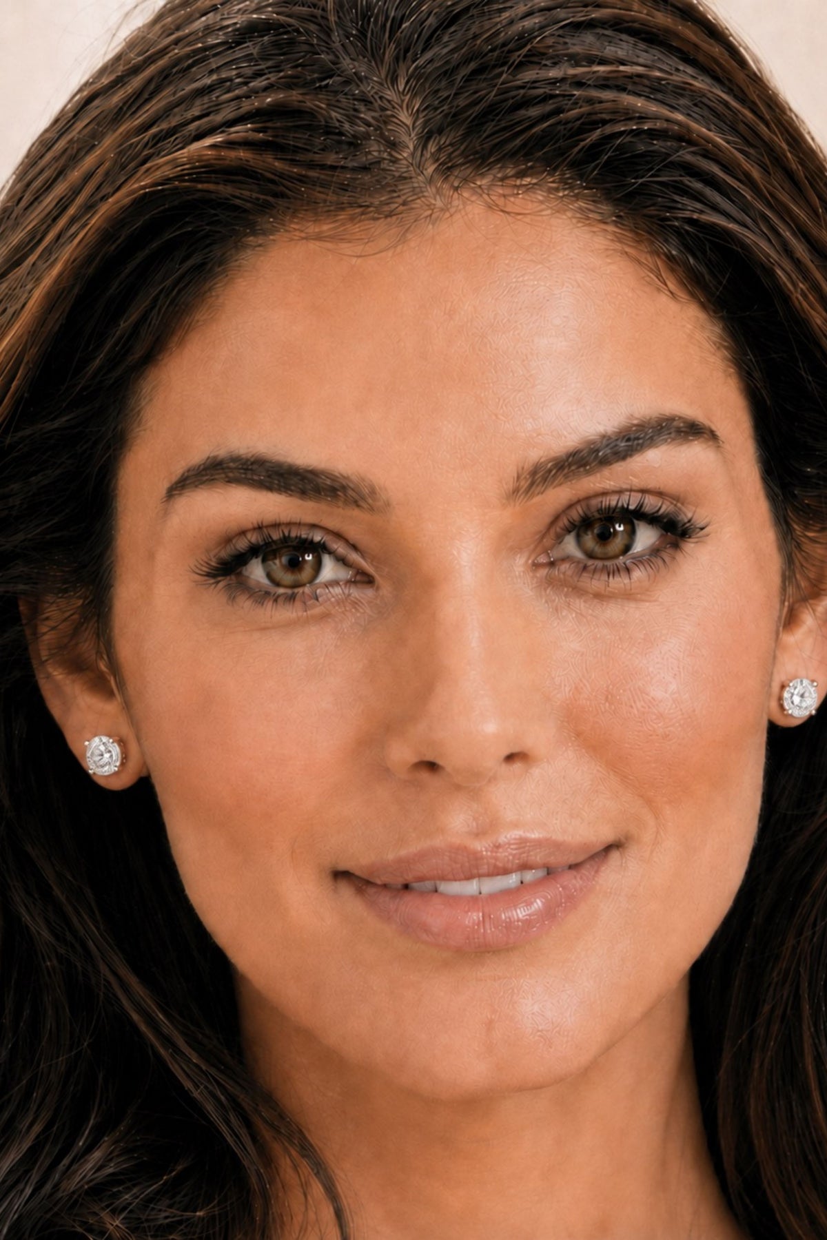A close-up portrait of a woman wearing small round brilliant-cut diamond stud earrings. The image is tightly cropped to show her face and hair, with a soft, warm neutral background.