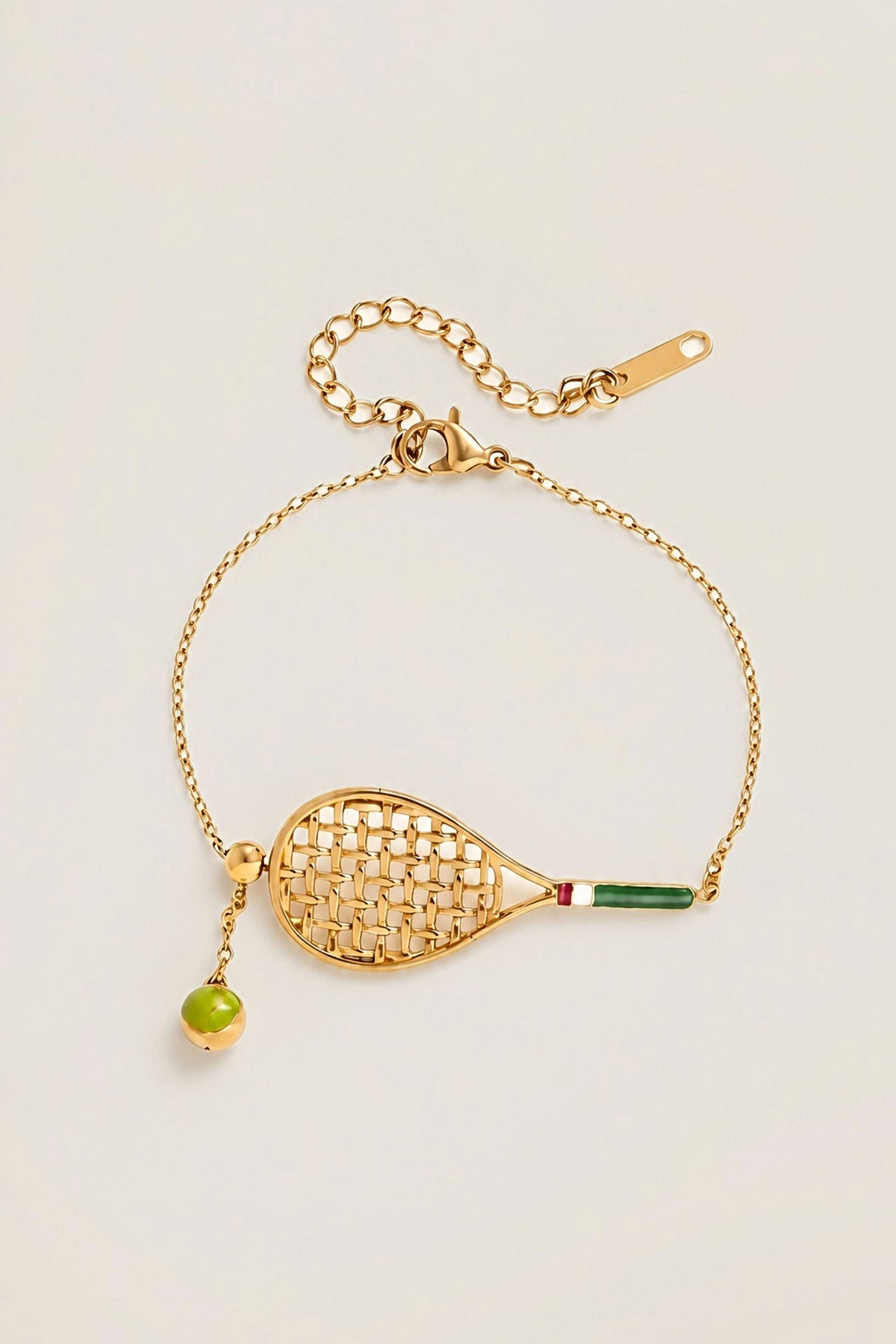 Country Club Tennis Bracelet in 14K gold PVD with an enamel tennis racket charm and a green enamel tennis ball, on a warm greige backdrop.
