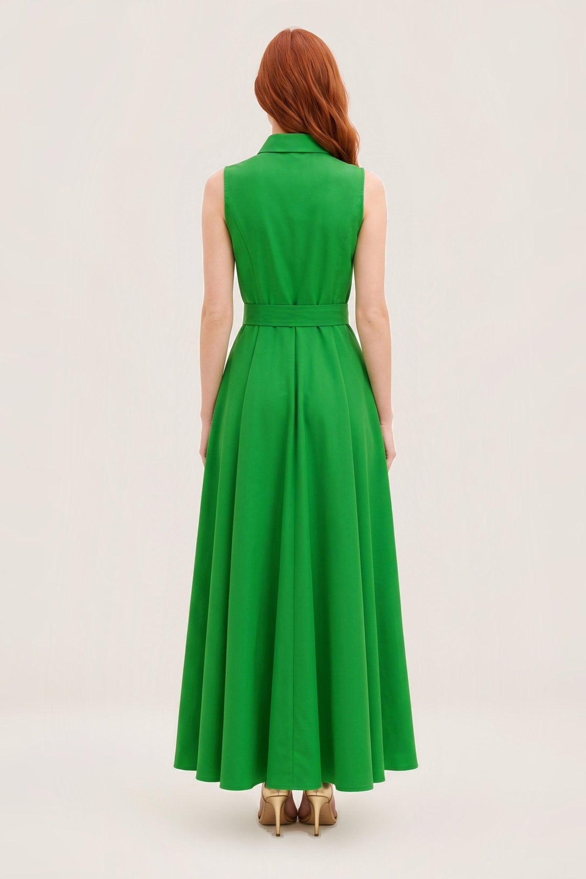 A woman with red hair is shown from the back wearing a sleeveless, high-neck, long-length green dress with a matching waist belt, paired with gold heels, against a plain off-white studio background.