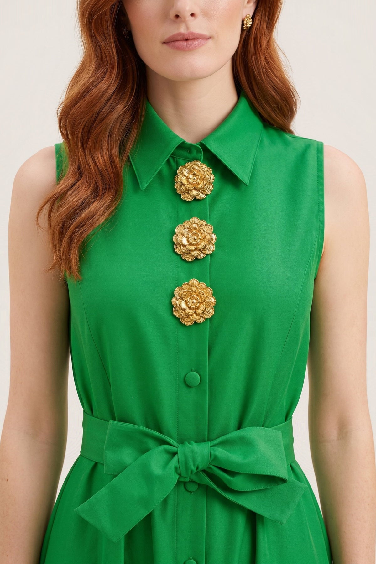 A close-up of a woman wearing a bright green sleeveless collared dress with three large gold flower-shaped buttons down the front and a matching waist tie, against a light beige background.