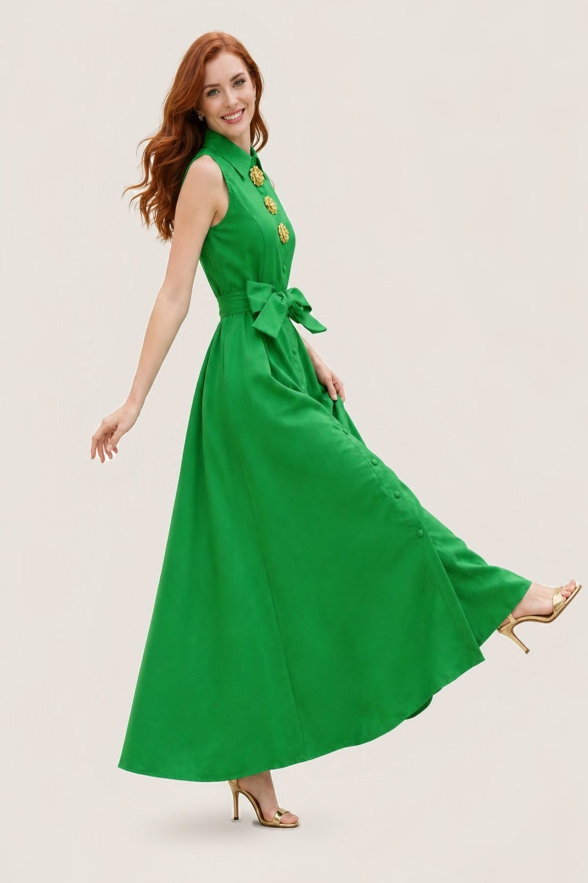 A woman mid-step in the Emerald Mikado belted A-line maxi dress, sleeveless with a pointed collar, gold rosette buttons and a bow belt, the full skirt swinging, on a warm neutral studio background.