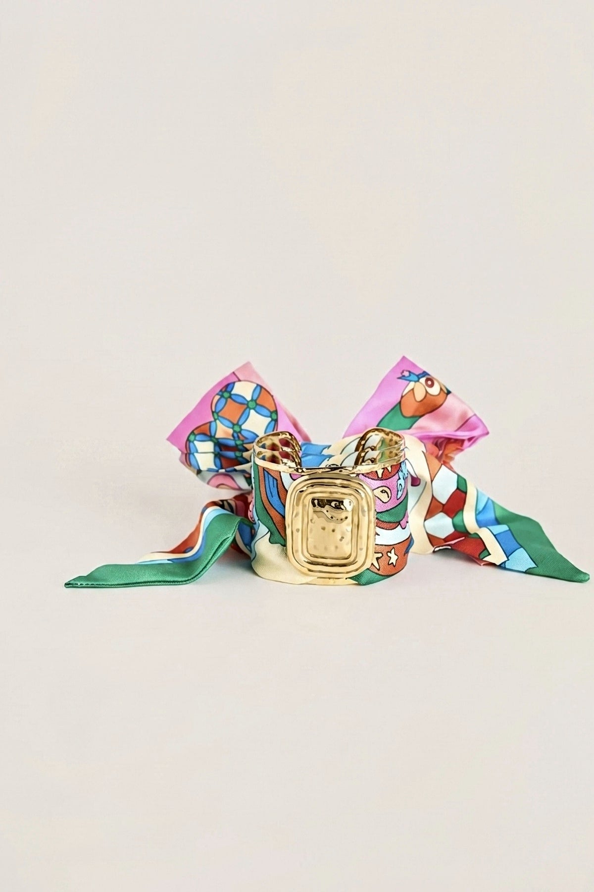 Evie Pink Multi Scarf Cuff, a gold cuff wrapped in a pink multicolour print scarf tied in a bow, on a warm greige backdrop.