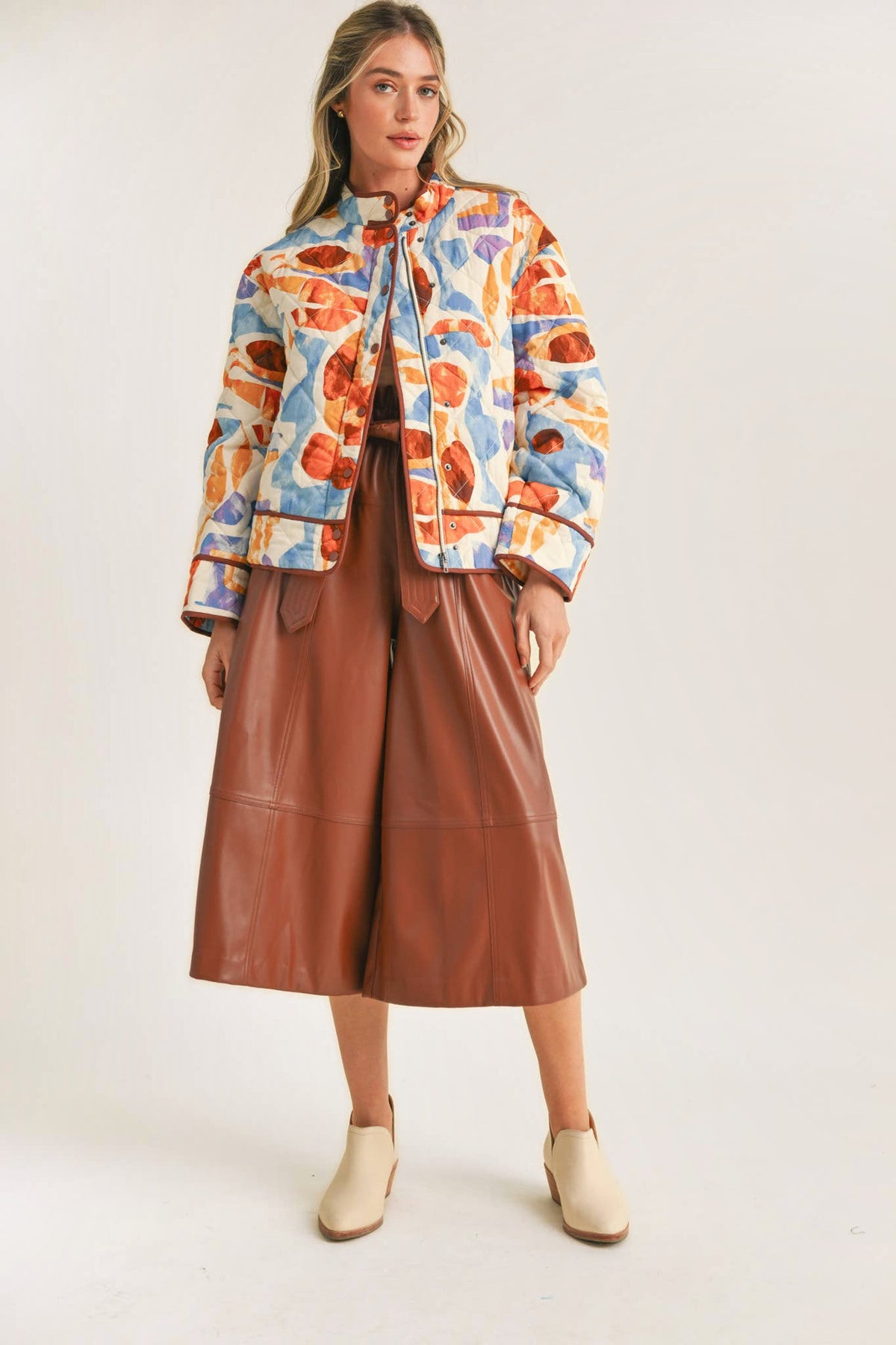 A woman modeling a colorful abstract print bomber jacket, paired with brown wide-leg faux-leather cropped pants and cream ankle boots, against a plain off-white studio background.
