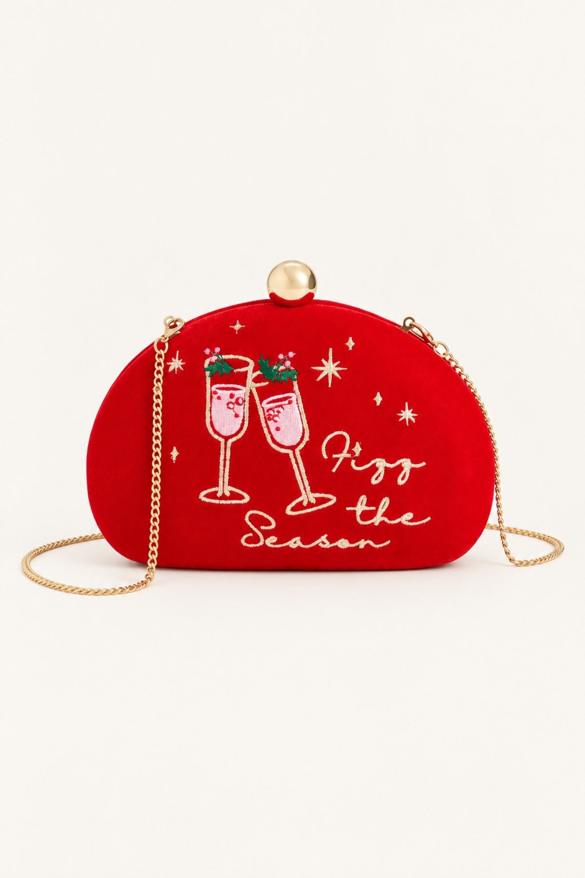 A red velvet crescent-shaped clutch bag with a gold-tone chain strap and top clasp. The front features white embroidered champagne glasses, festive starbursts, and the text 'Fizz the Season'.