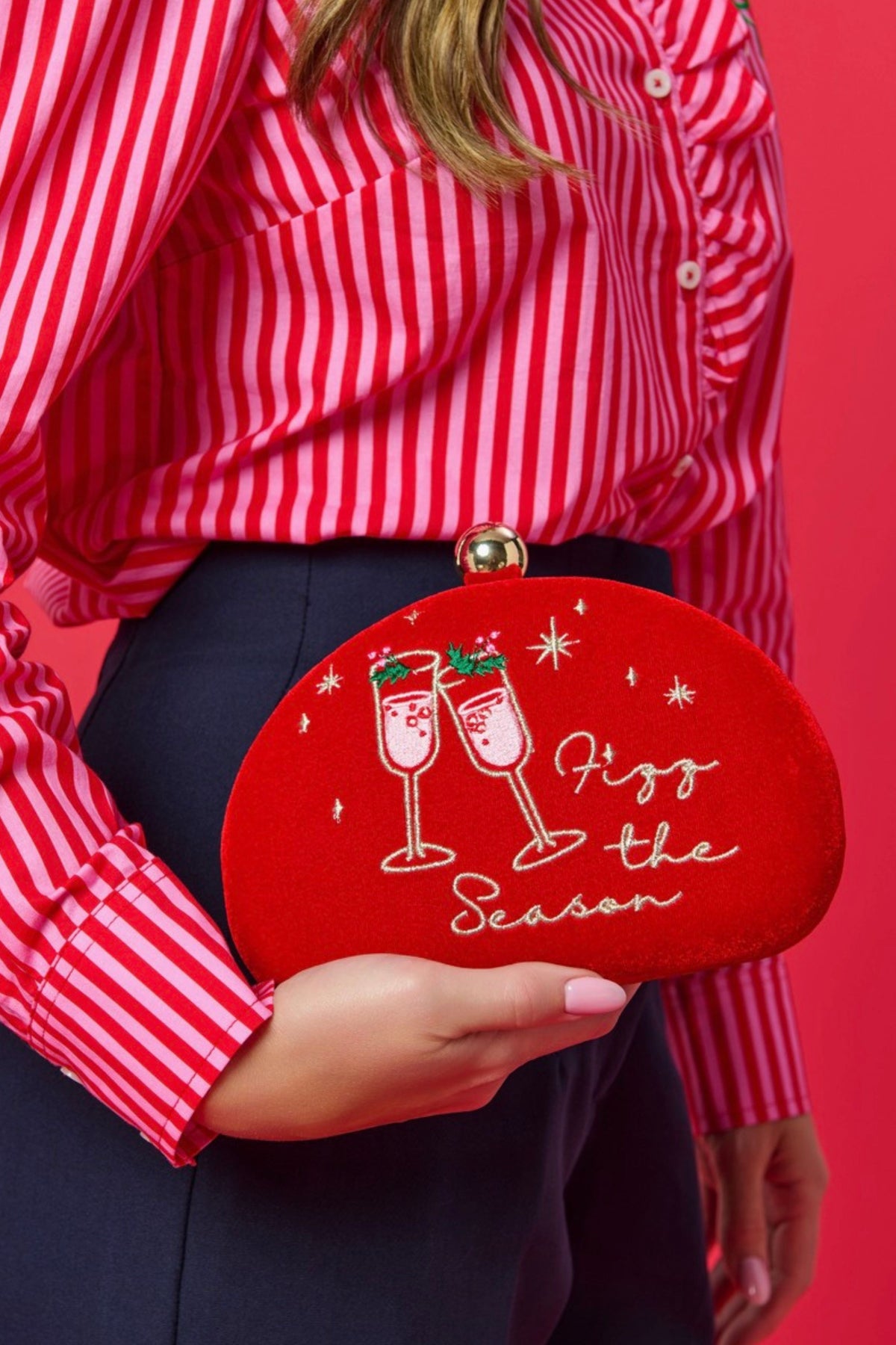 A person in a red and white striped shirt holds a red oval clutch bag with white script reading 'Fizz the Season' and two champagne glasses printed on the front, against a solid red background.