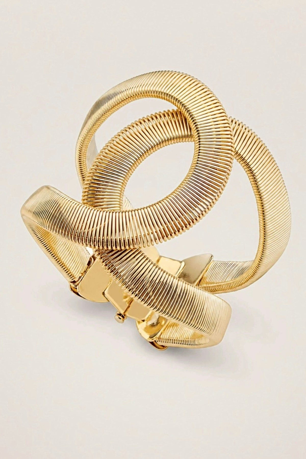 Giselle Bracelet, a sculptural gold cuff of two textured interlocking curves with a hinge closure, on a warm greige backdrop.