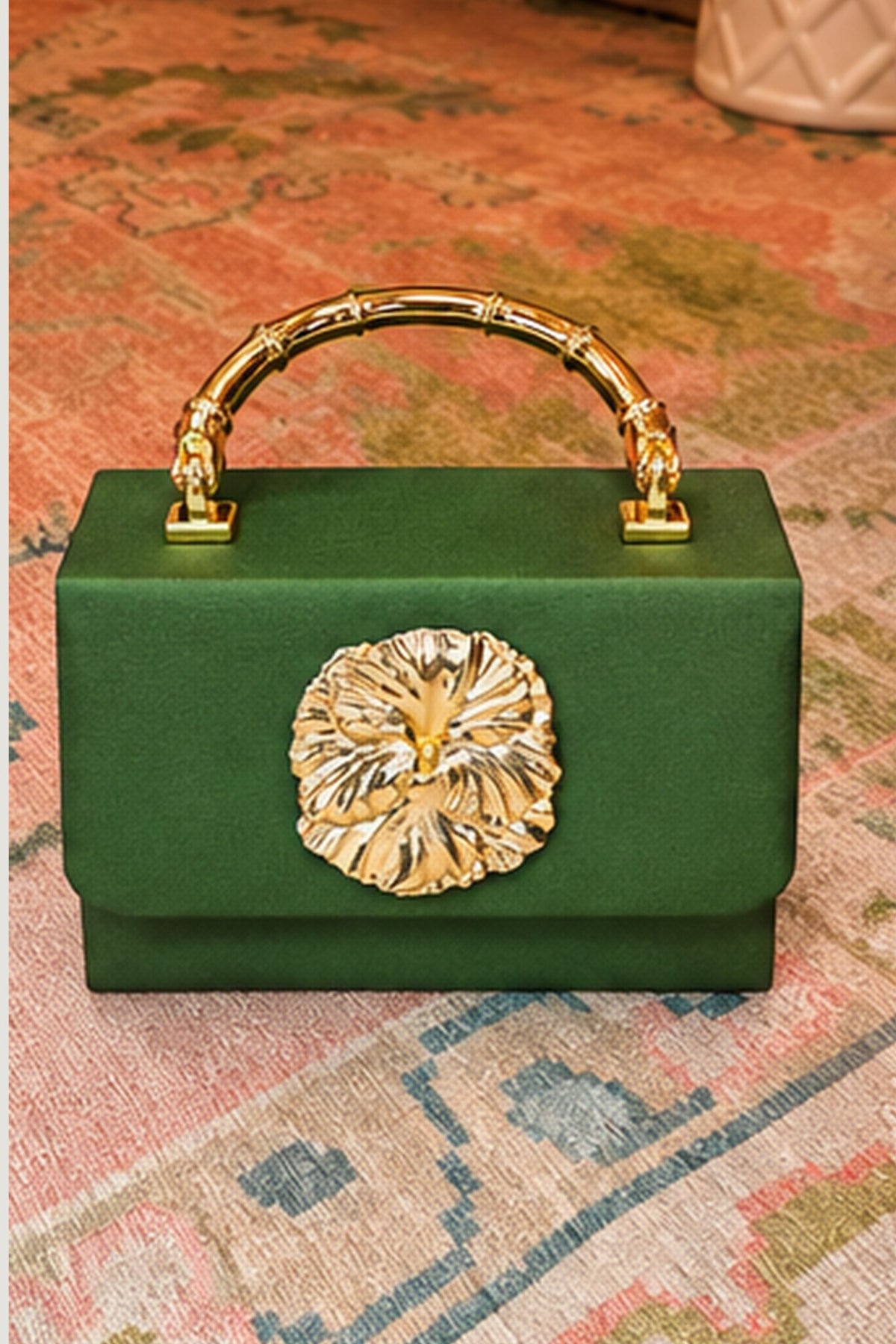 A structured green handbag with a bamboo-style gold handle and a large gold flower clasp on the front, photographed on a patterned rug.