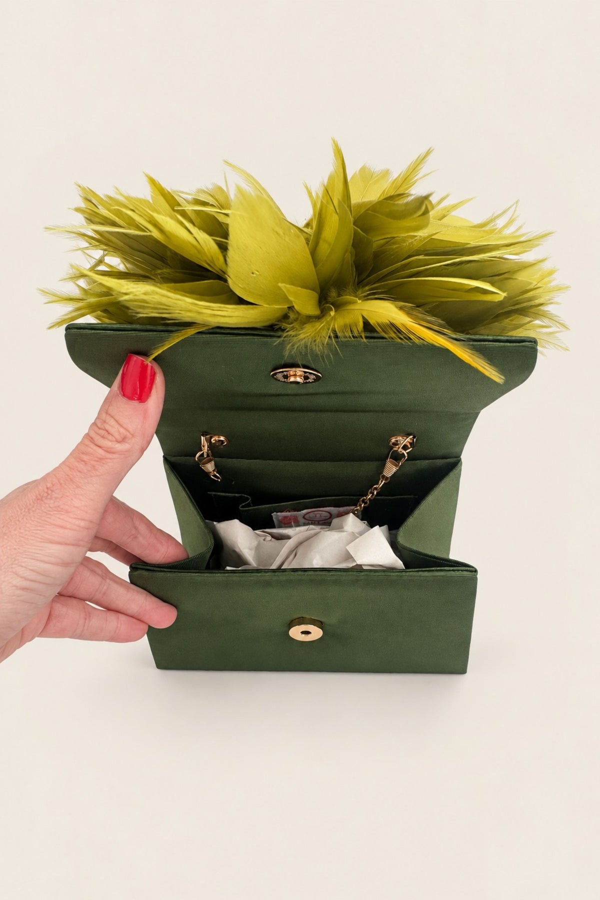 A dark green structured handbag with gold-tone hardware, shown open with a hand holding the left side. A bright lime-green artificial flower arrangement is placed on top of the bag, and a white tissue or paper insert is visible inside the compartments.