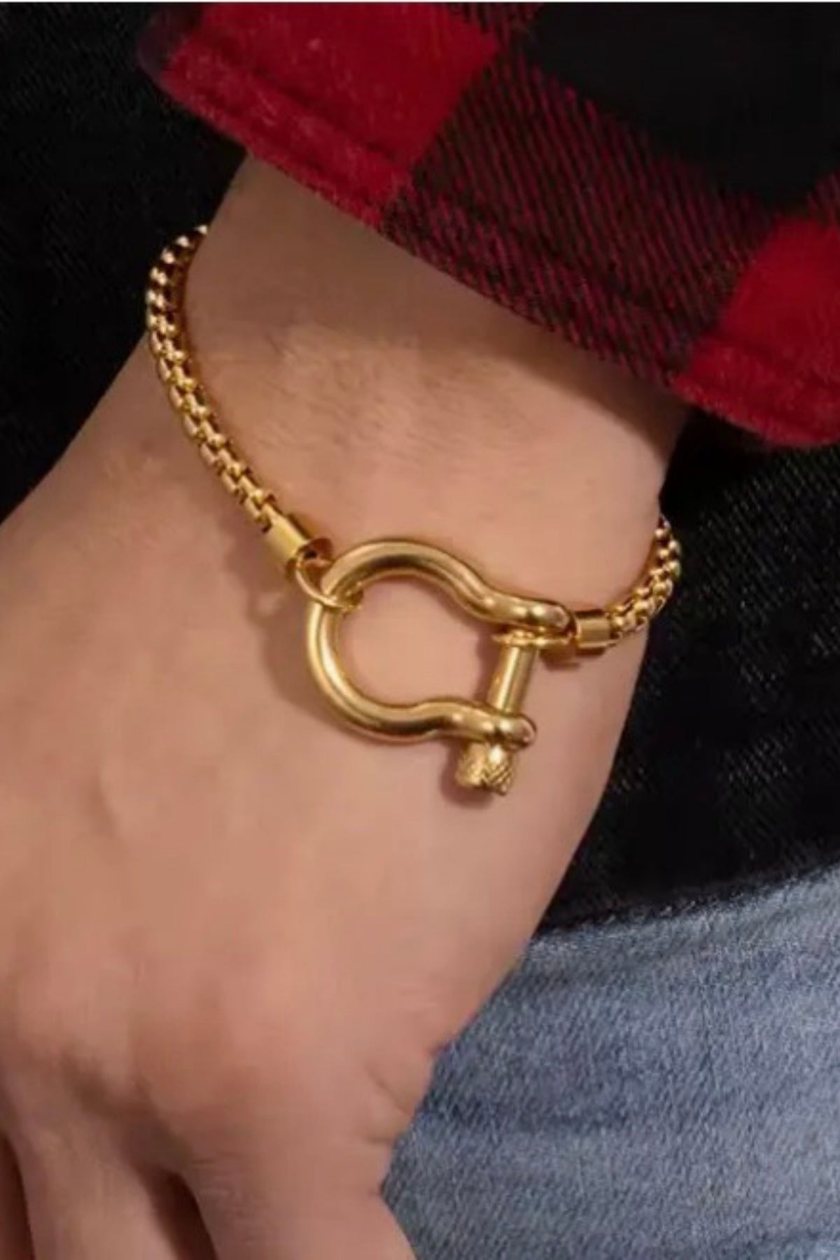 A gold-tone chain bracelet with a large circular ring and shackle-like clasp, shown worn on a person's wrist. The person is wearing a red and black plaid cuff or sleeve, with dark and light clothing visible in the background.