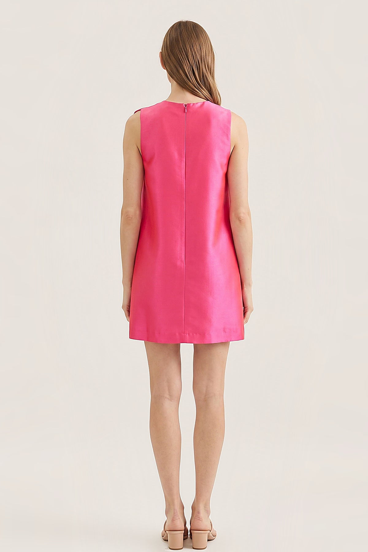 A woman viewed from the back wearing a sleeveless bright pink shift dress with a straight neckline and a back zipper, paired with beige heeled sandals against a plain light beige studio background.