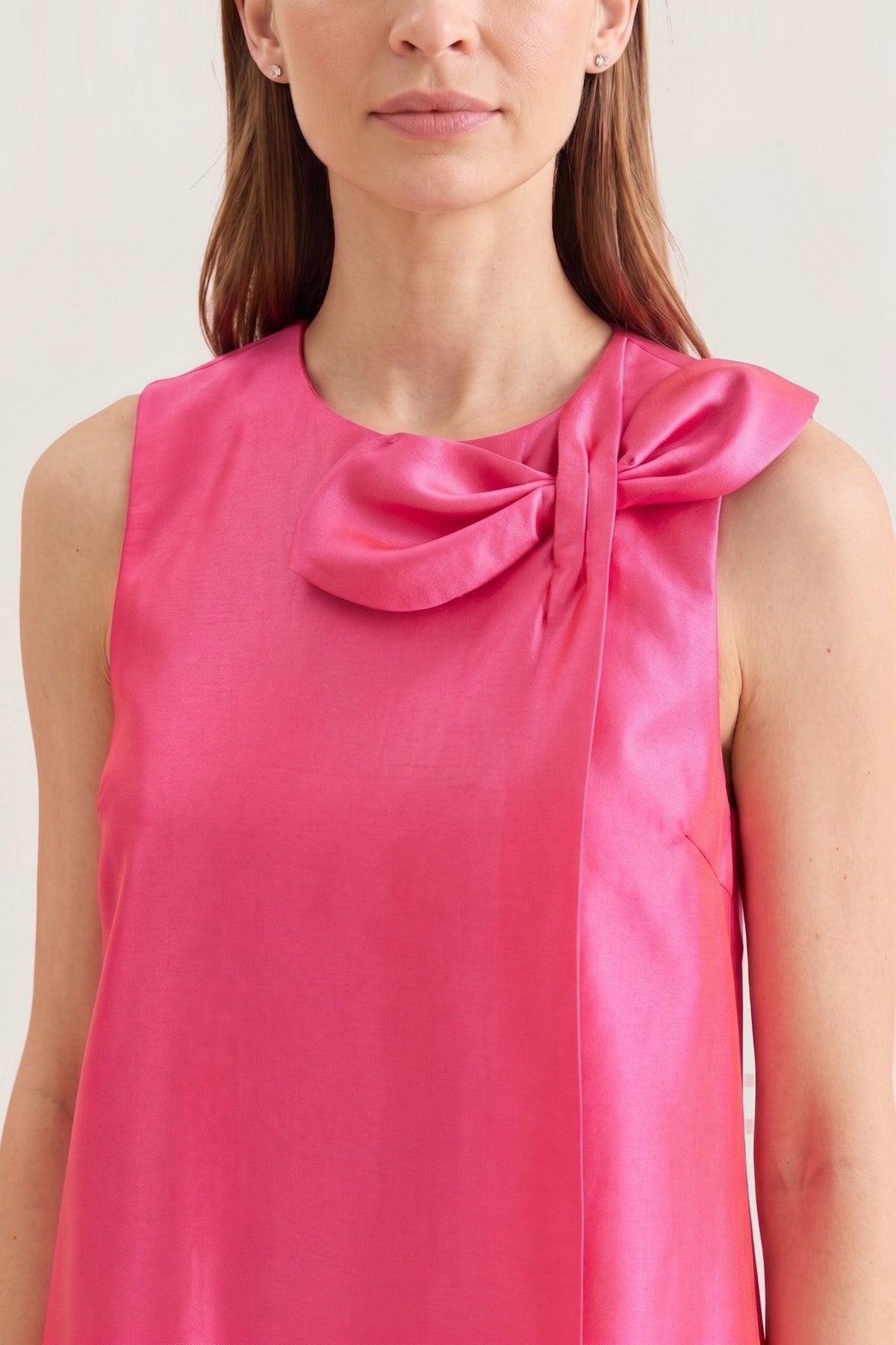 A close-up of a woman wearing a sleeveless bright pink satin top with a large bow detail at the neckline, set against a plain light beige background.