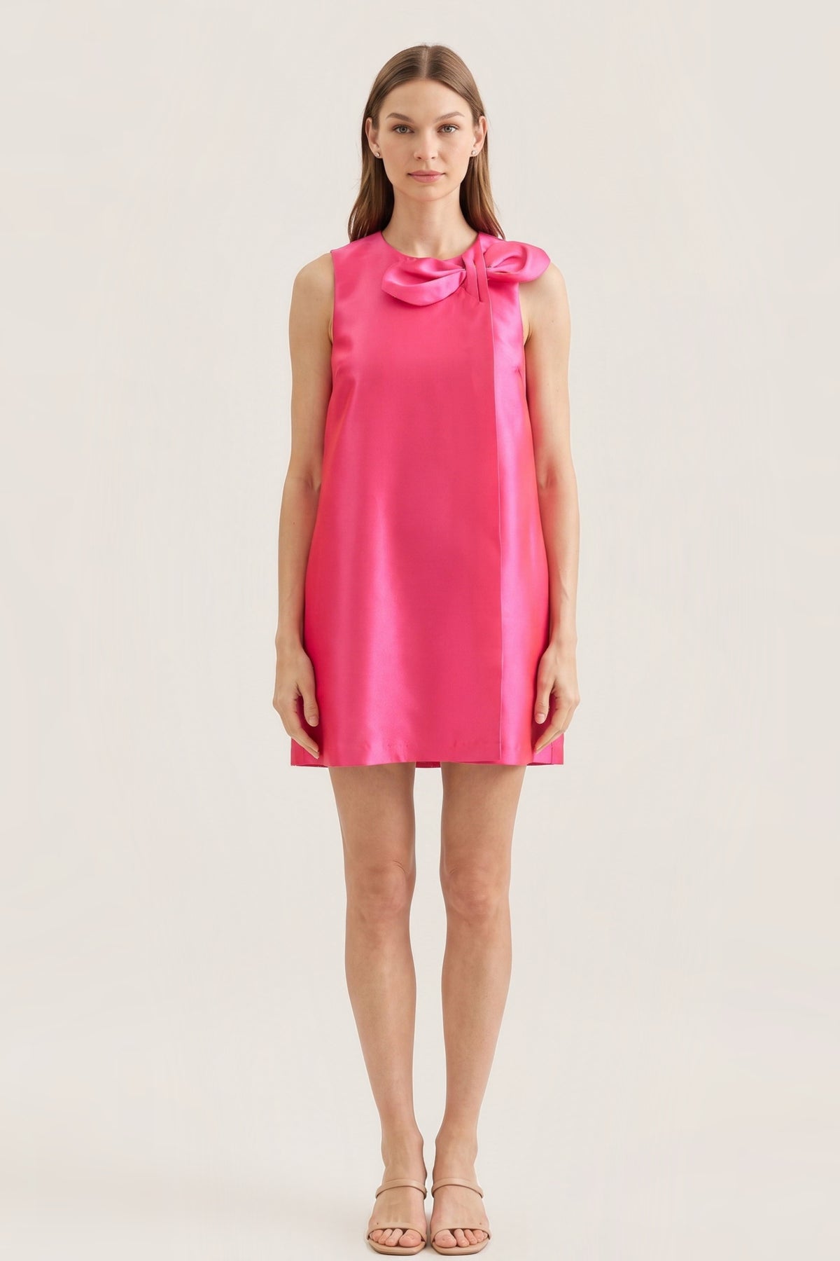 A woman modeling a sleeveless hot pink satin mini dress with a large bow detail at the neckline, set against a plain light beige studio background.