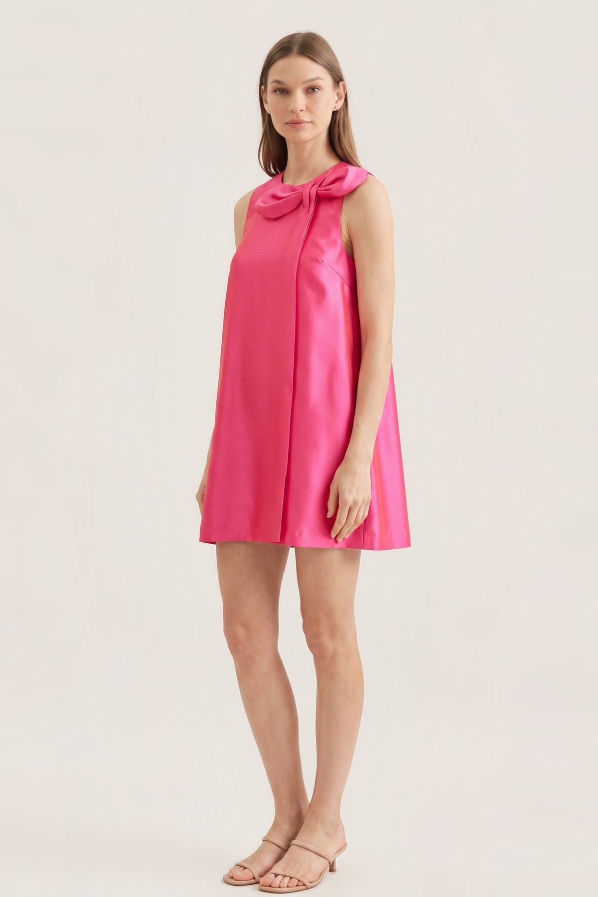 A woman modeling a sleeveless hot pink mini dress with a large bow detail at the neckline, photographed against a plain light beige studio background.