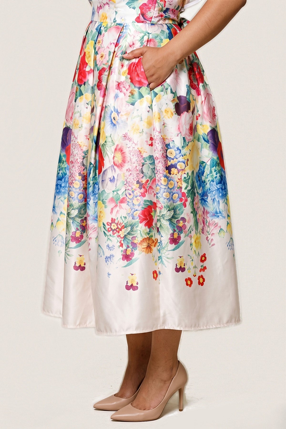 Close-up of the Jardin Bloom Dress skirt: satin pleats from the waist, a hand in the hidden side pocket, and the floral print fading into a wide ivory hem border.