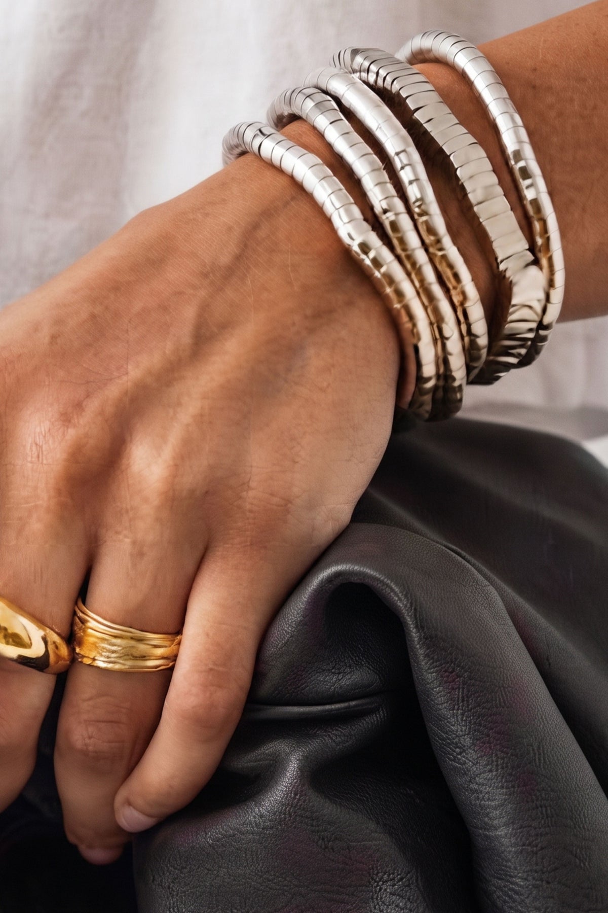 A close-up of a person's hand wearing multiple textured silver bangle bracelets and two gold rings, resting on a dark leather surface.