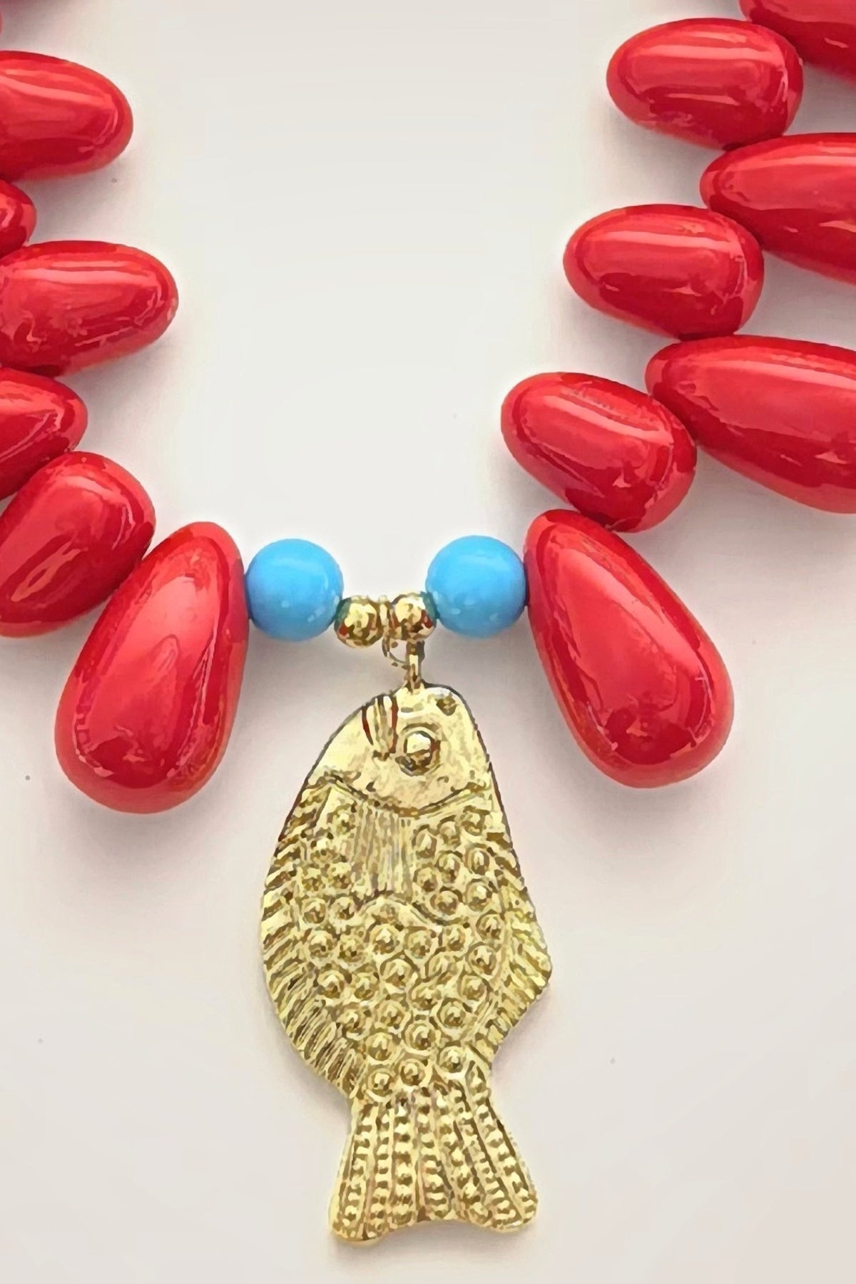 Close-up of the hammered gold fish pendant hanging between two turquoise beads on the red Lucky Fish resin bead necklace.