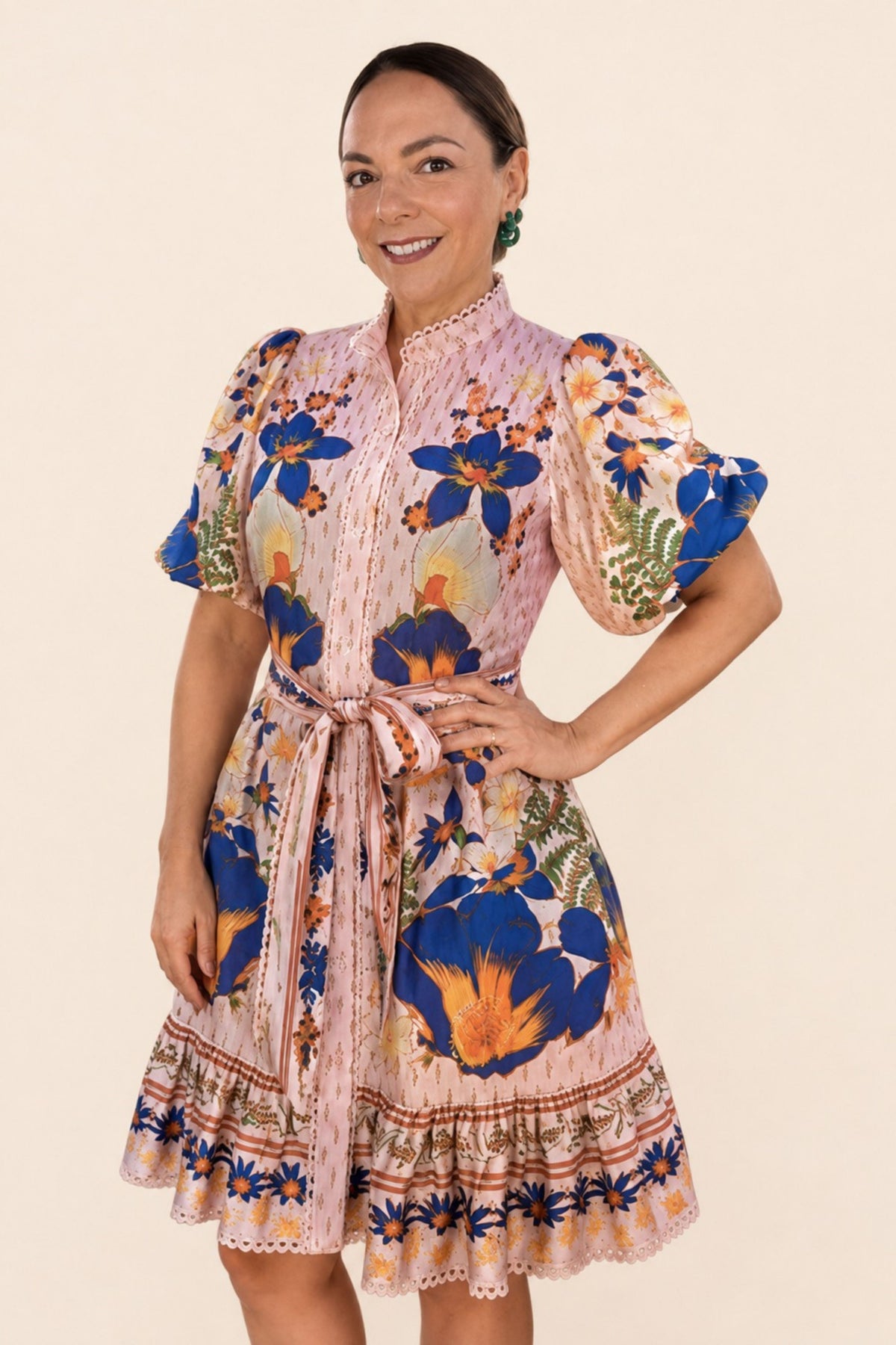 A woman modeling a peach floral print dress with blue, orange, and green botanical motifs, featuring puff sleeves, a high collar, and a matching waist tie, set against a solid beige background.