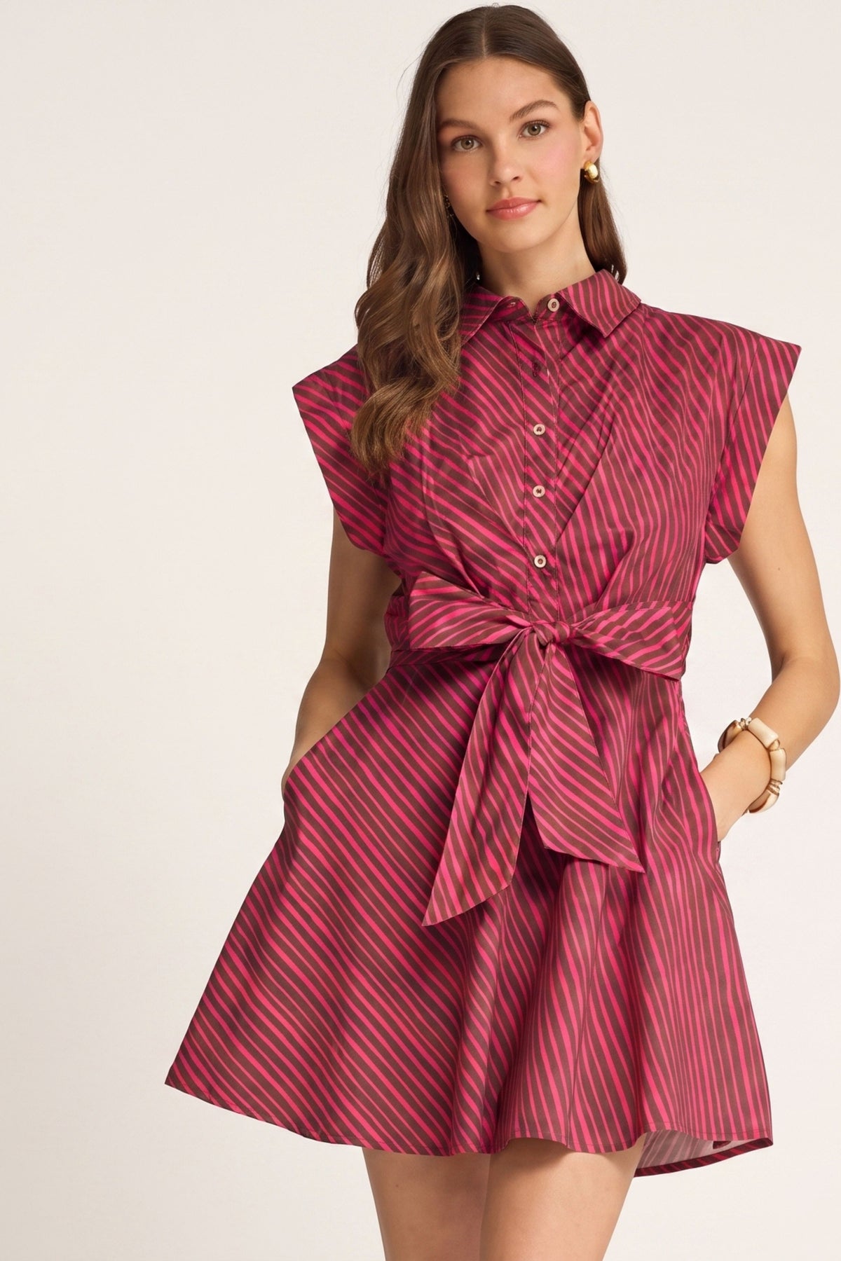 A woman models a cap sleeve collared shirt dress with a tie waist and a raspberry and brown diagonal stripe pattern, set against a plain light background.