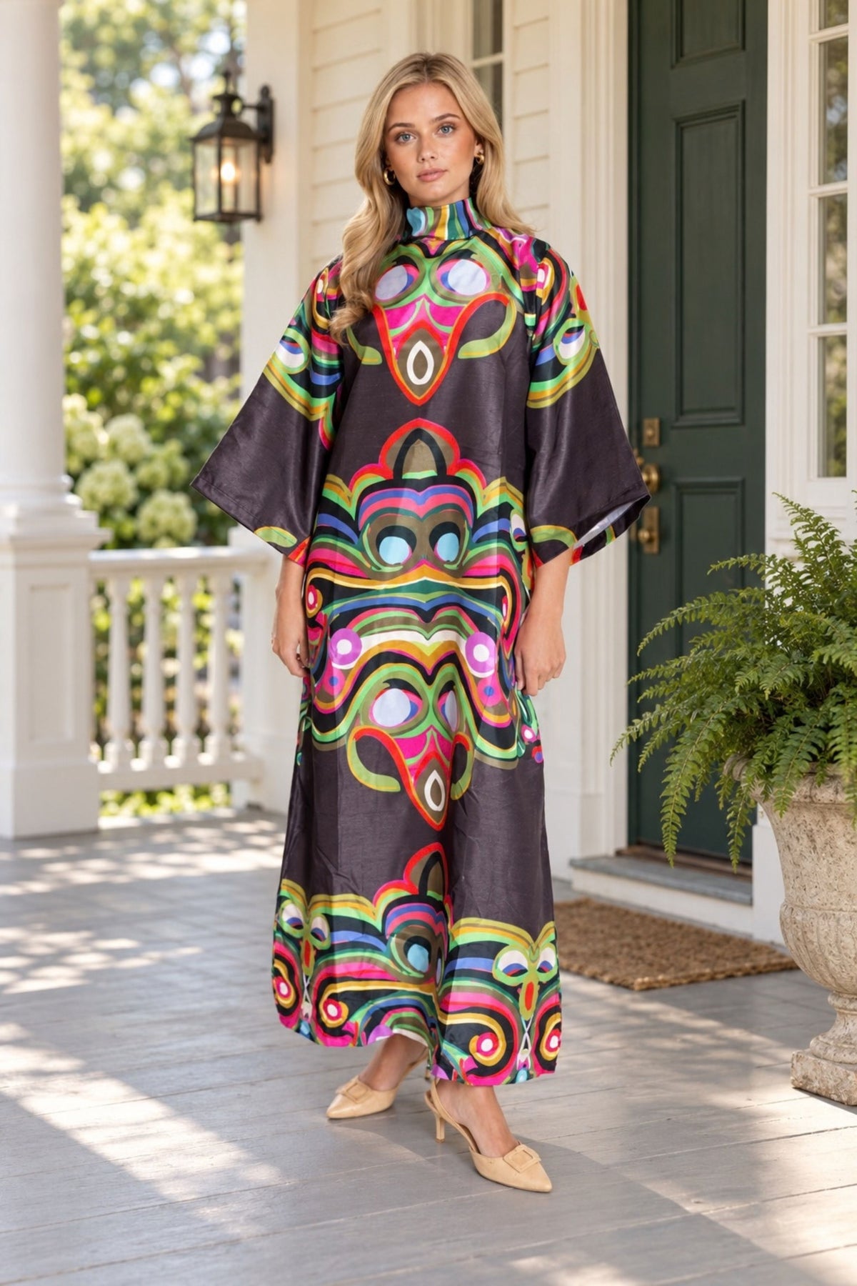 A woman stands on a porch wearing a long, loose-fitting dress with wide sleeves and a bold psychedelic swirl pattern in pink, green, yellow, blue, and orange on a dark base. The background shows a white house porch with a green door and potted plants.